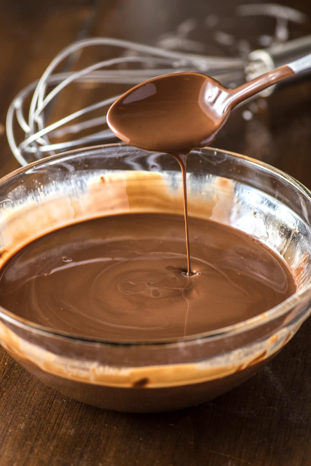 Clear glass mixing bowl full of melted chocolate with a teaspoon of melted chocolate held above and dripping into the bowl. There is a whisk in the background.