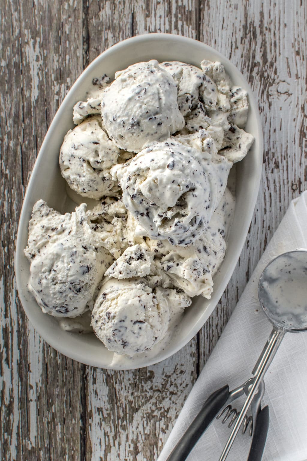 Oval, white bowl piled high with vanilla, ice cream full of shaved chocolate. On a rustic barn board back drop with a folded napkin at the side on which rests an empty ice cream scoop.