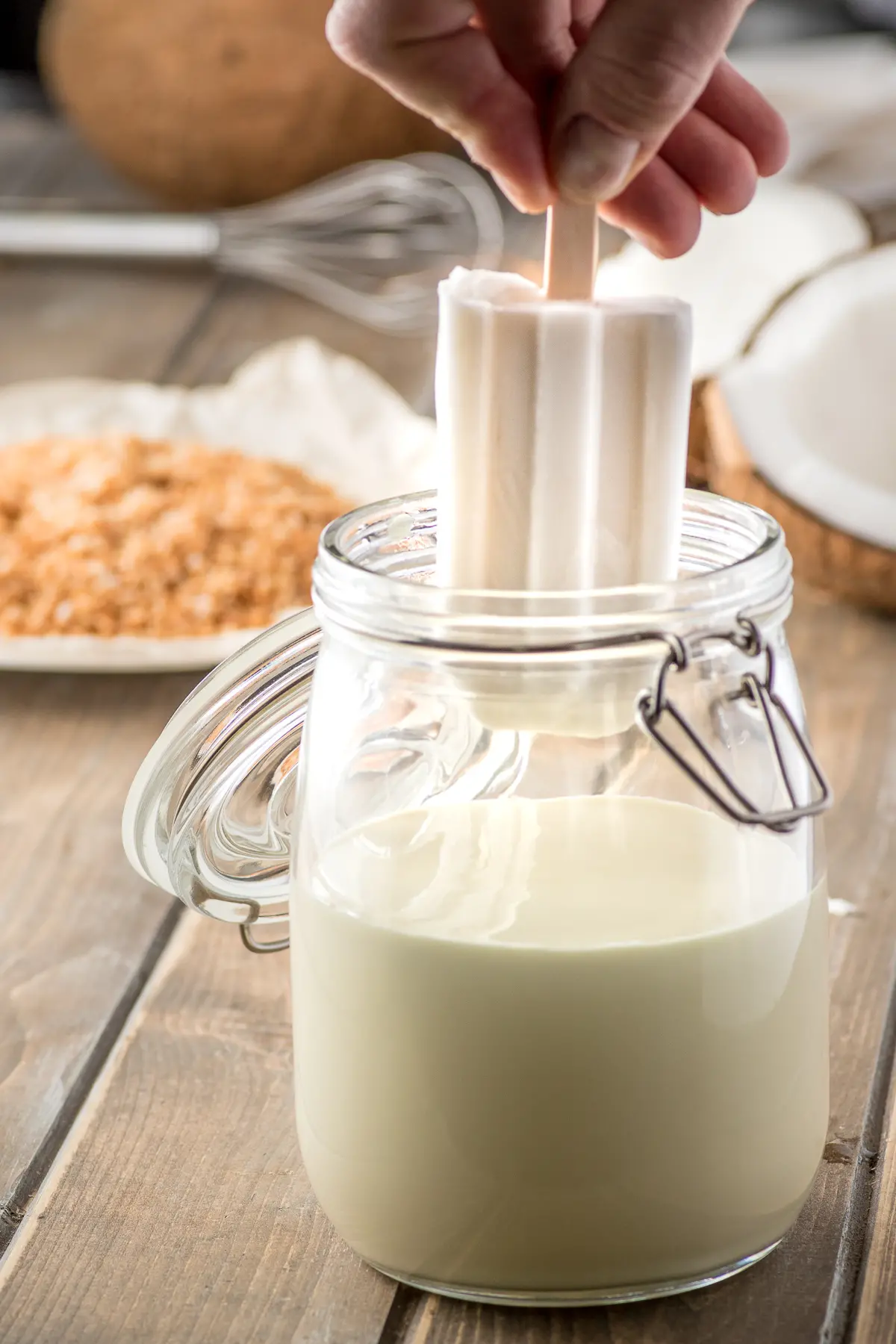 a glass jar filled with white chocolate coating with a white coconut popsicle being dipped 