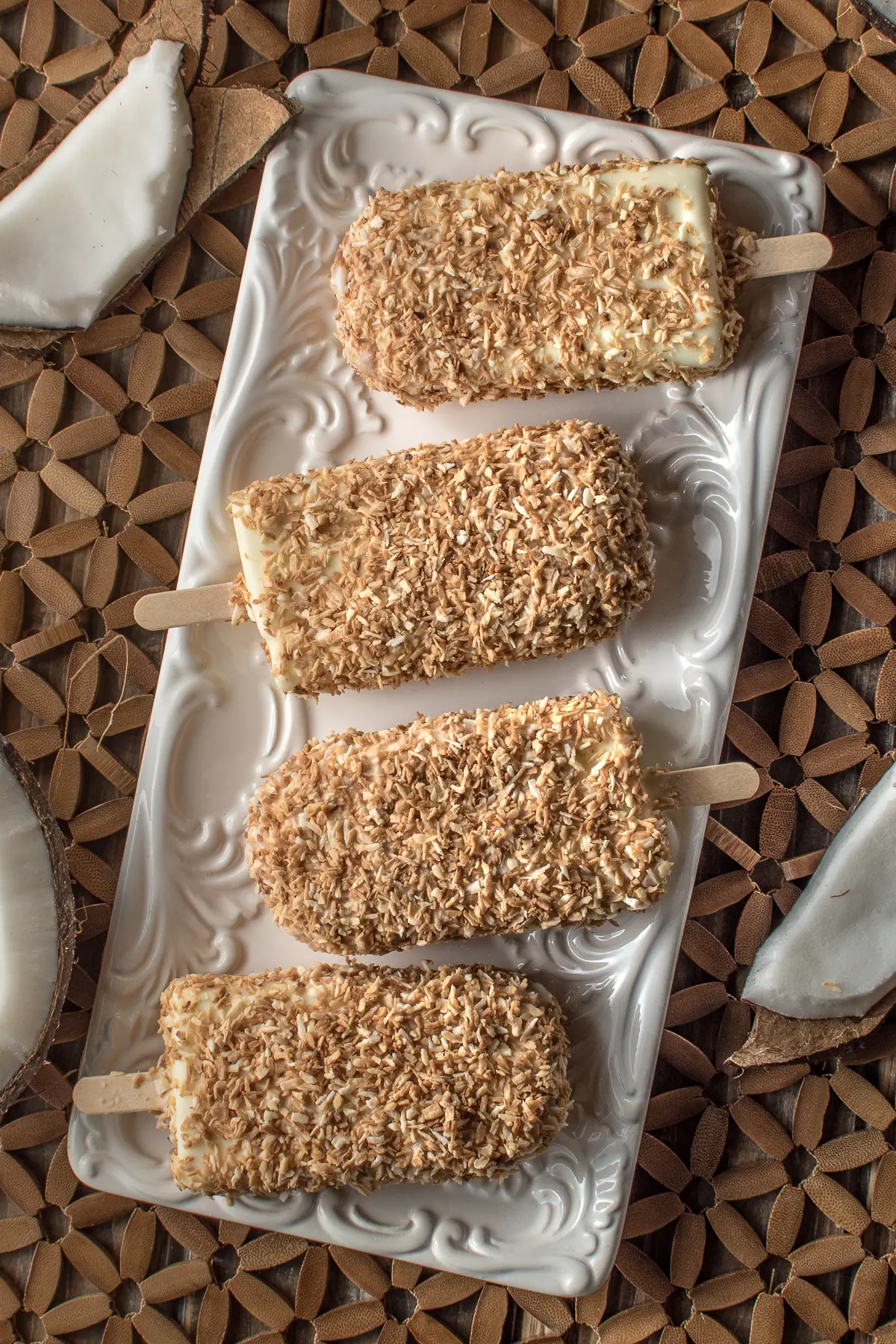 overhead shot of creamy coconut ice cream bars coated in toasted coconut resting on a white plate on a textured wood background