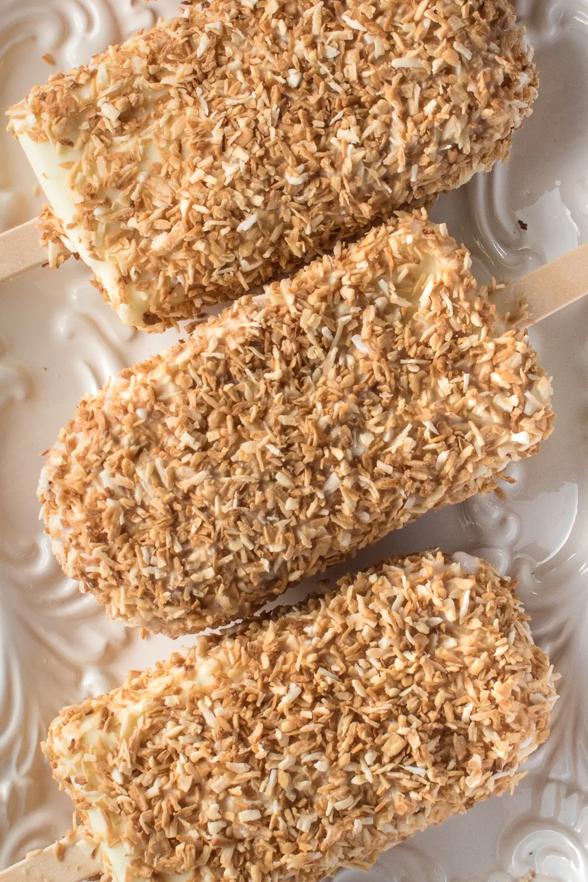homemade coconut ice cream bars coated in toasted coconut resting on a white textured plate. 