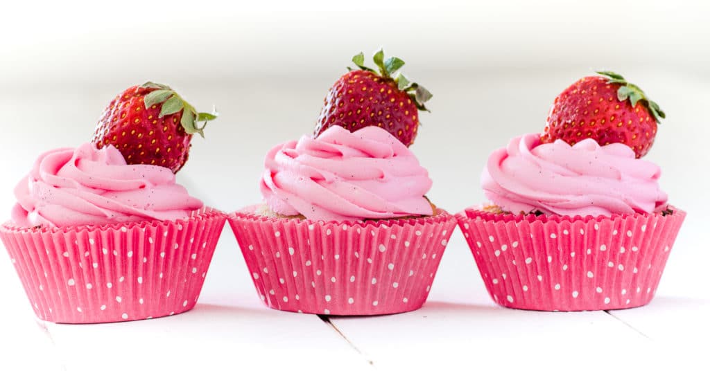 A row of 3 pink cupcakes topped with red ripe strawberries 