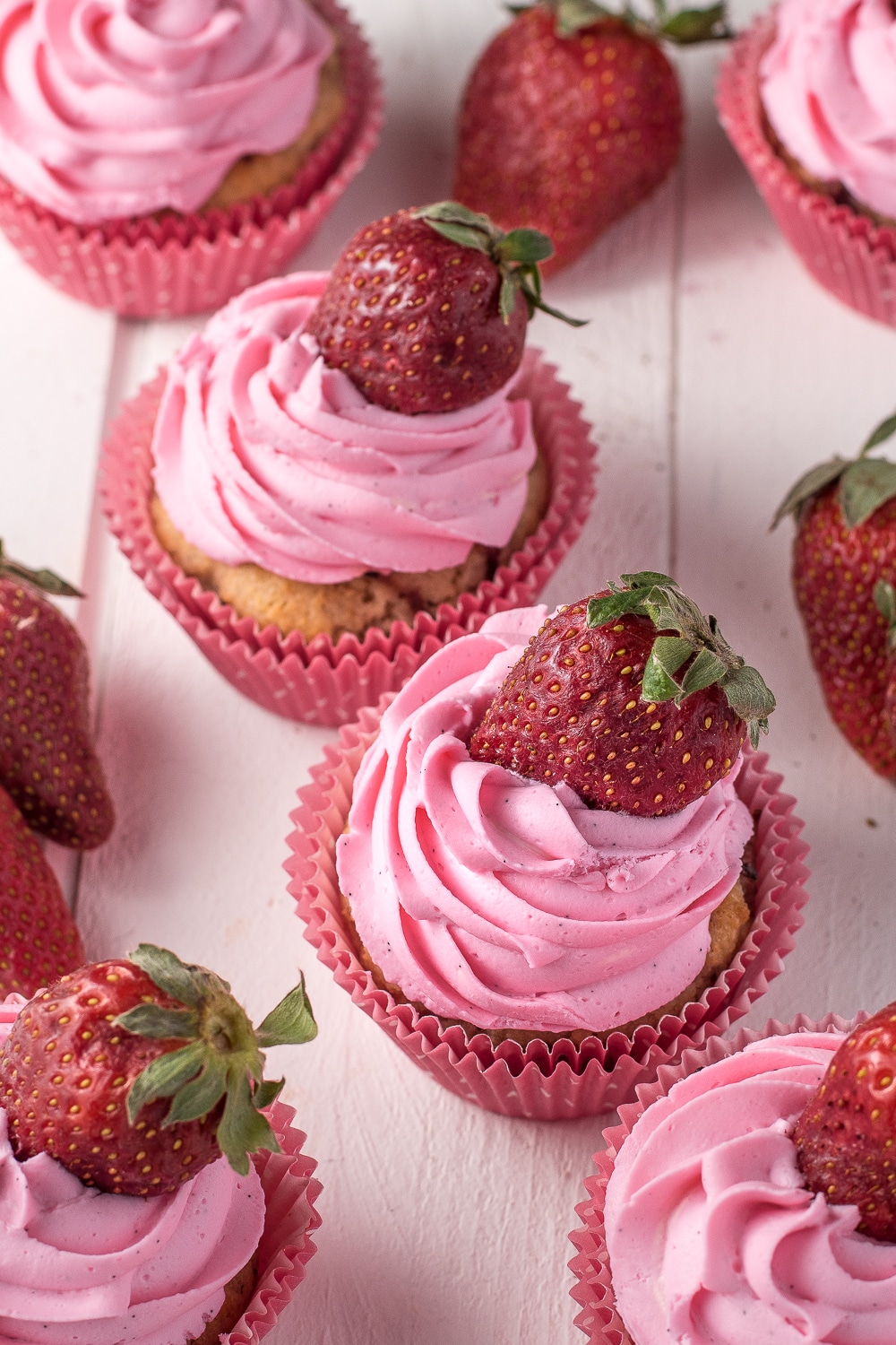 Pink icing topped strawberry cupcakes on a white wooden board topped with bright red fresh strawberries. 