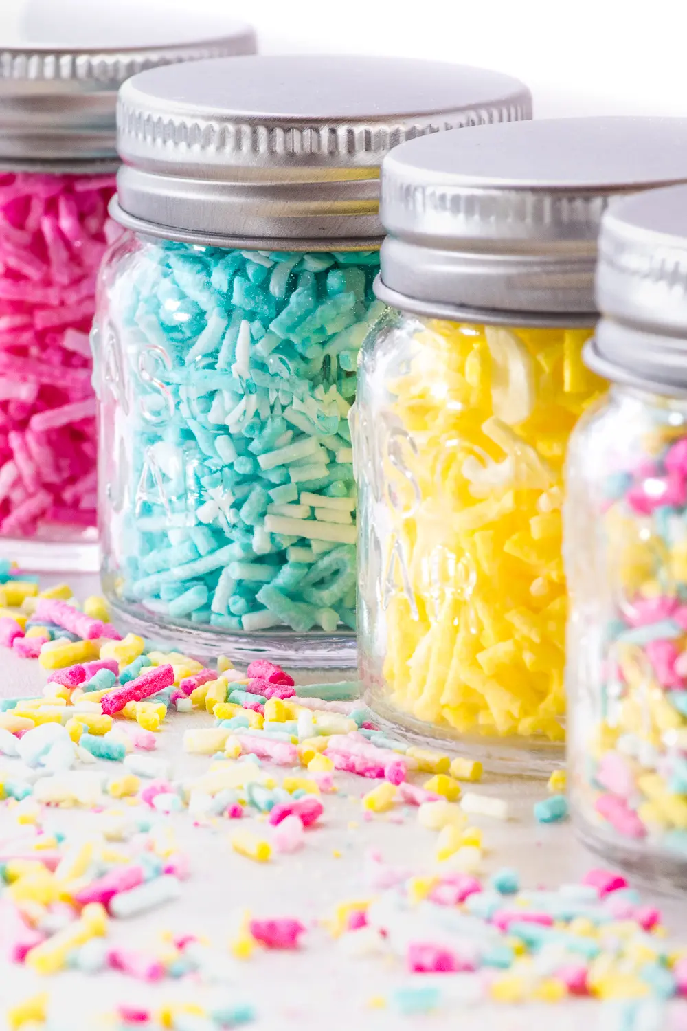 Small glass jars filled with colorful sprinkles.  Sprinkles are also coating the bright white tabletop. 