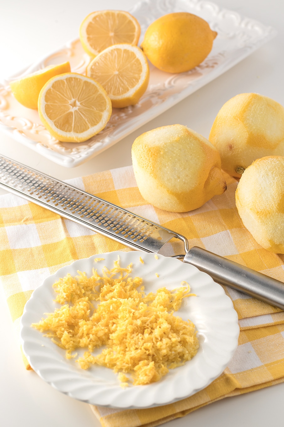 a plate of lemon zest, a long metal zester, with sliced and zested lemons on a white background
