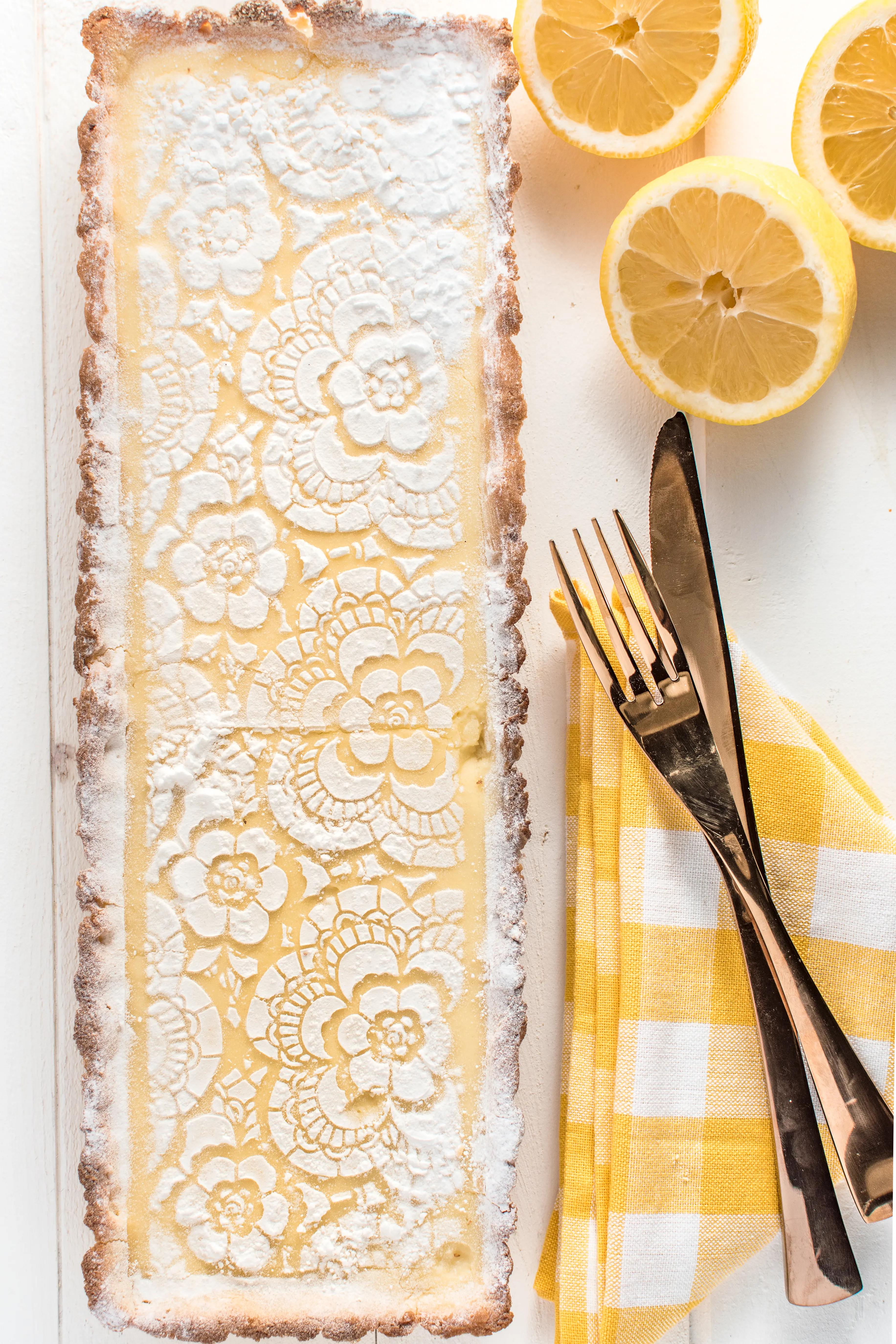 a top down photo of a rectangular lemon tart, with a gingham yellow napkin and fresh lemons on a white wooden background