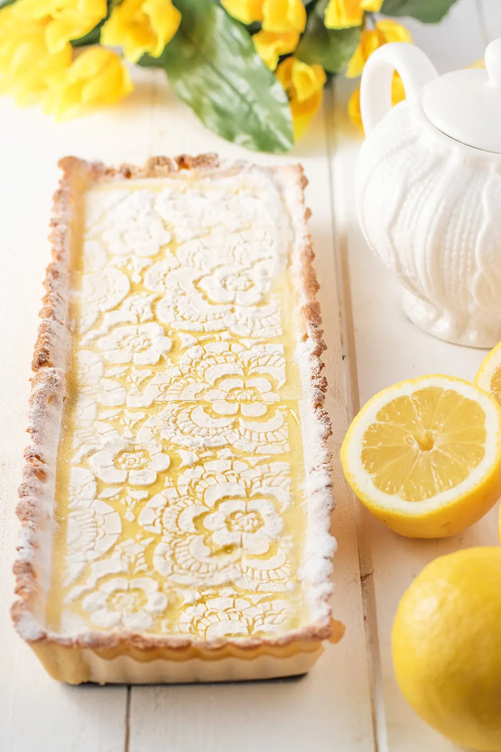 A freshly backed lemon cream tart, with yellow flowers, fresh lemons, and a pot of tea.
