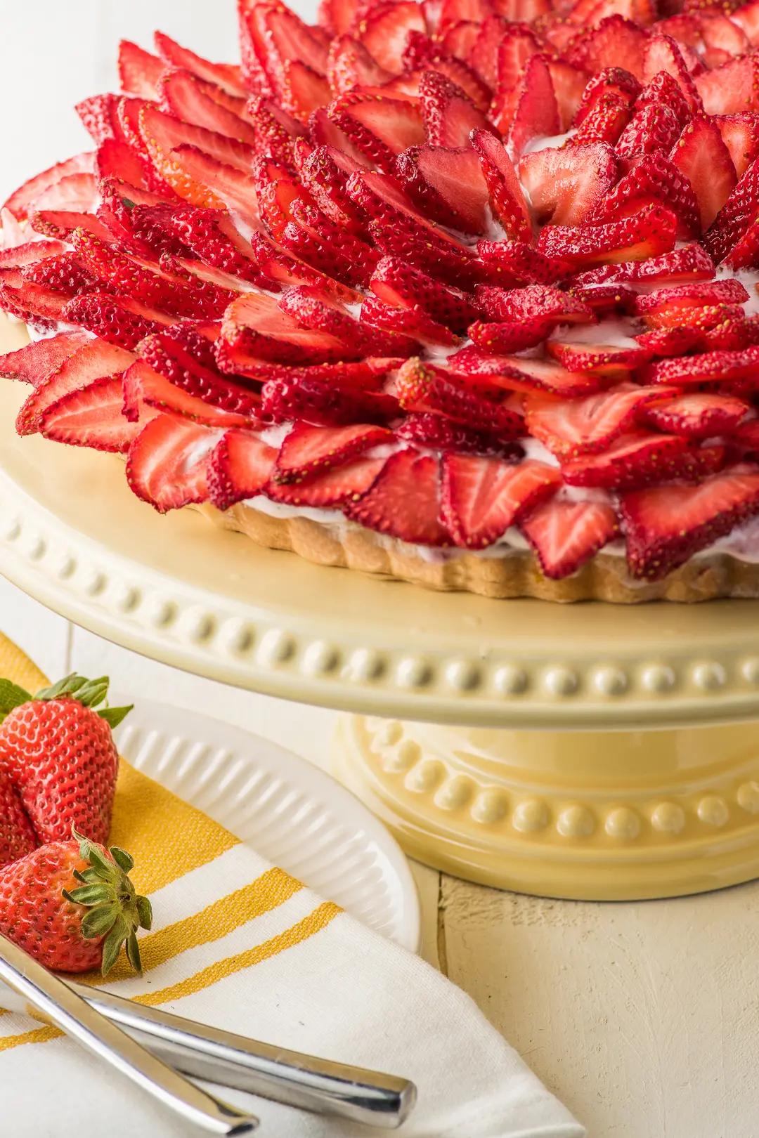 A strawberry cream pie topped with sliced strawberries on a yellow cake stand