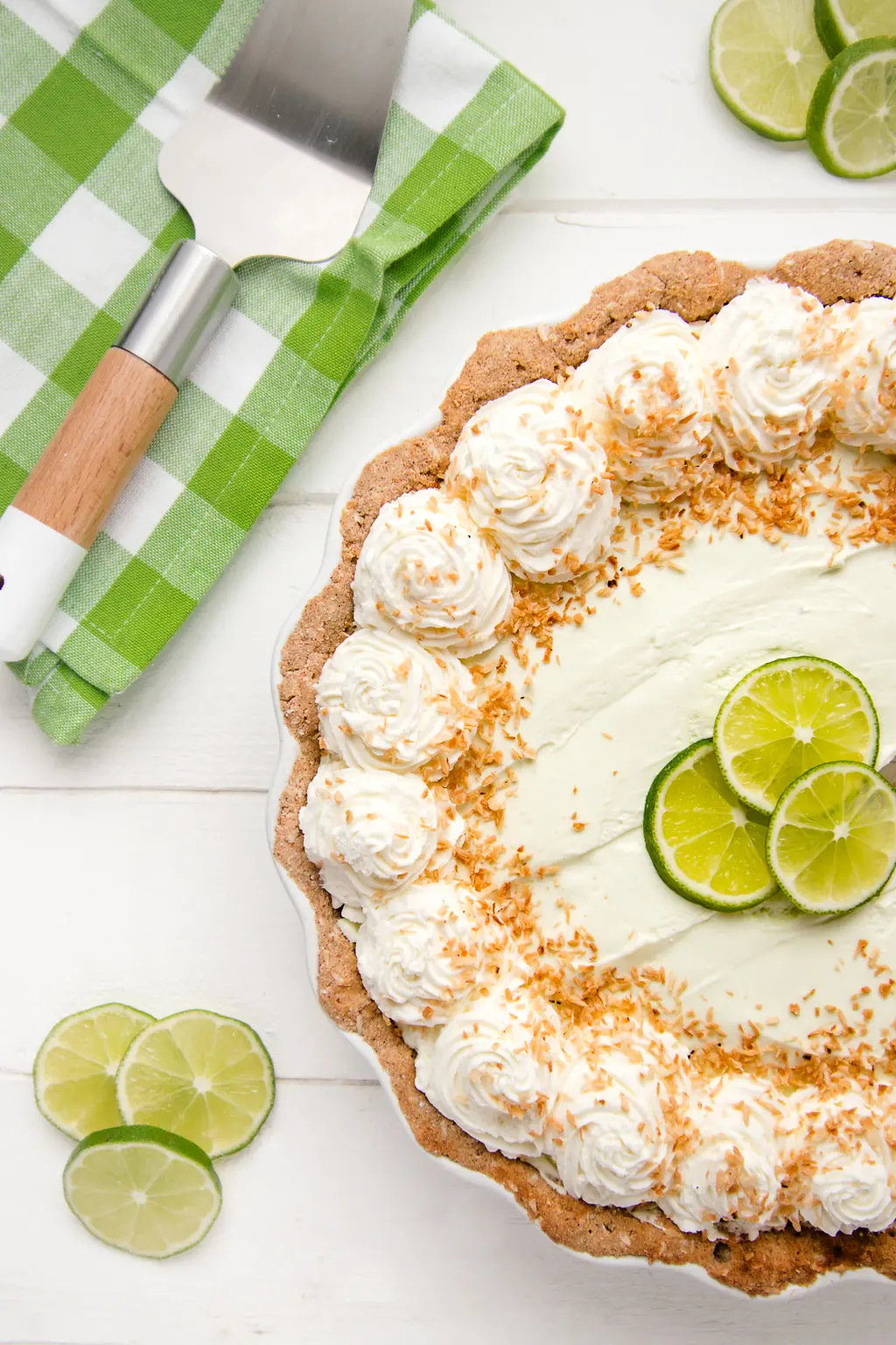A low carb key lime pie with whipped cream and toasted coconut on a white back ground with a green gingham napkin and sliced limes
