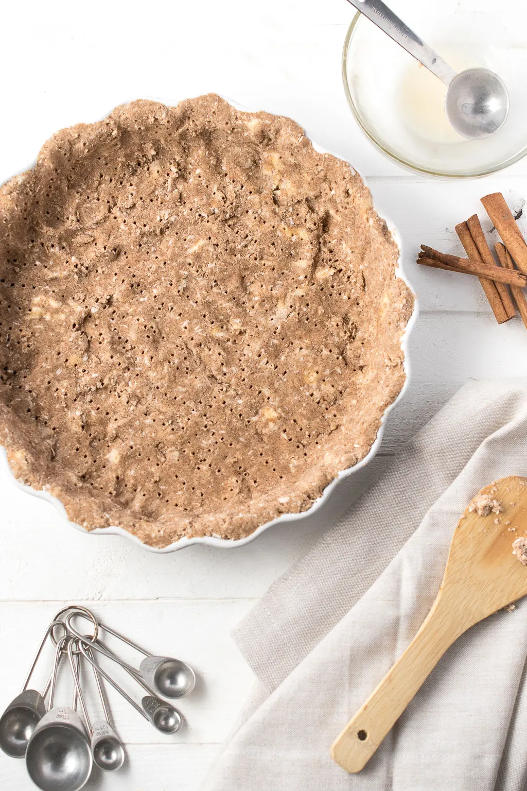 Baked keto pie crust in a pan on a white background, surrounded by cinnamon sticks, measuring spoons, and a wooden spoon which is resting on a napkin.