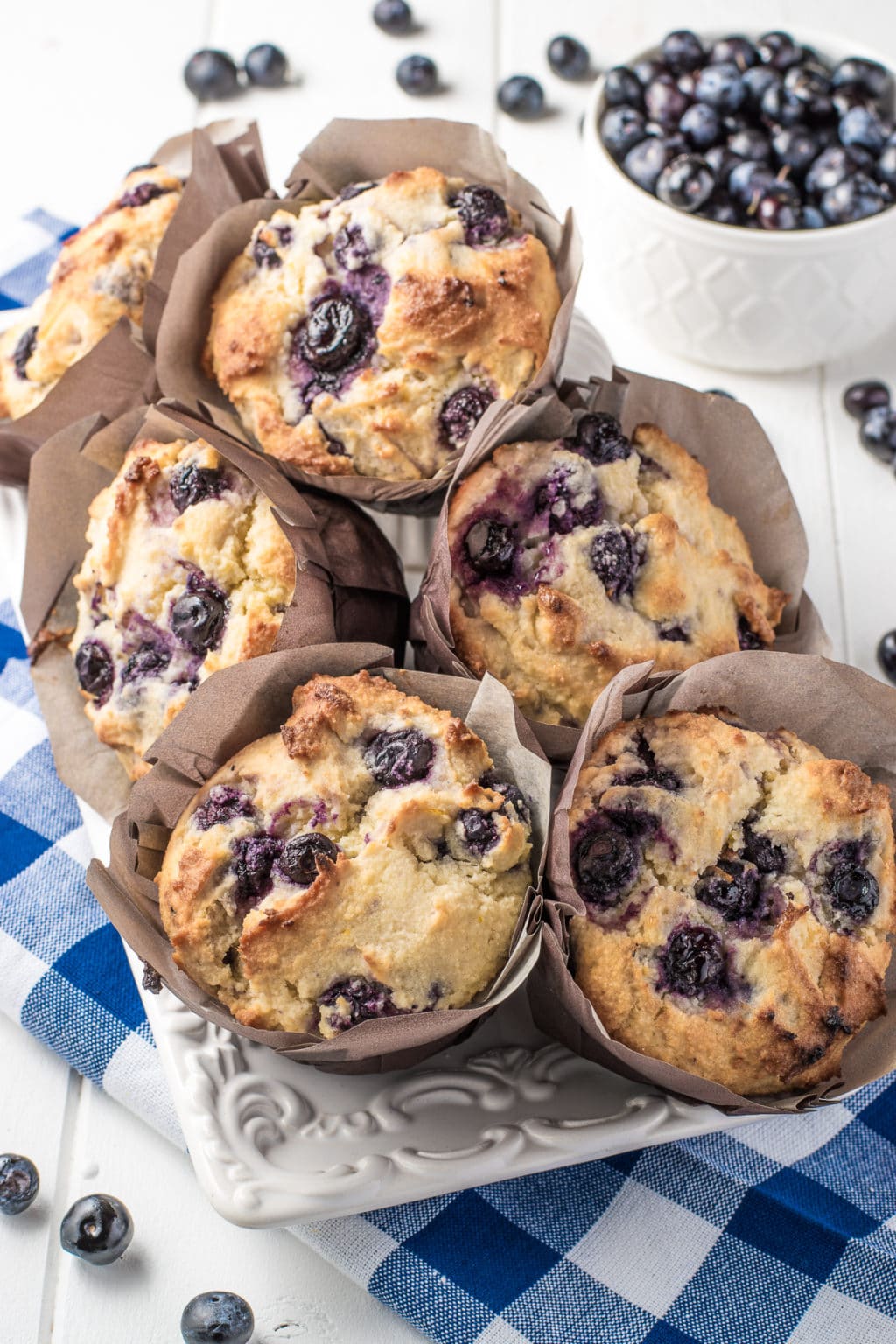 A stack of fresh homemade almond flour blueberry muffins on a white texture plate with a bright blue gingham napkin underneath. 