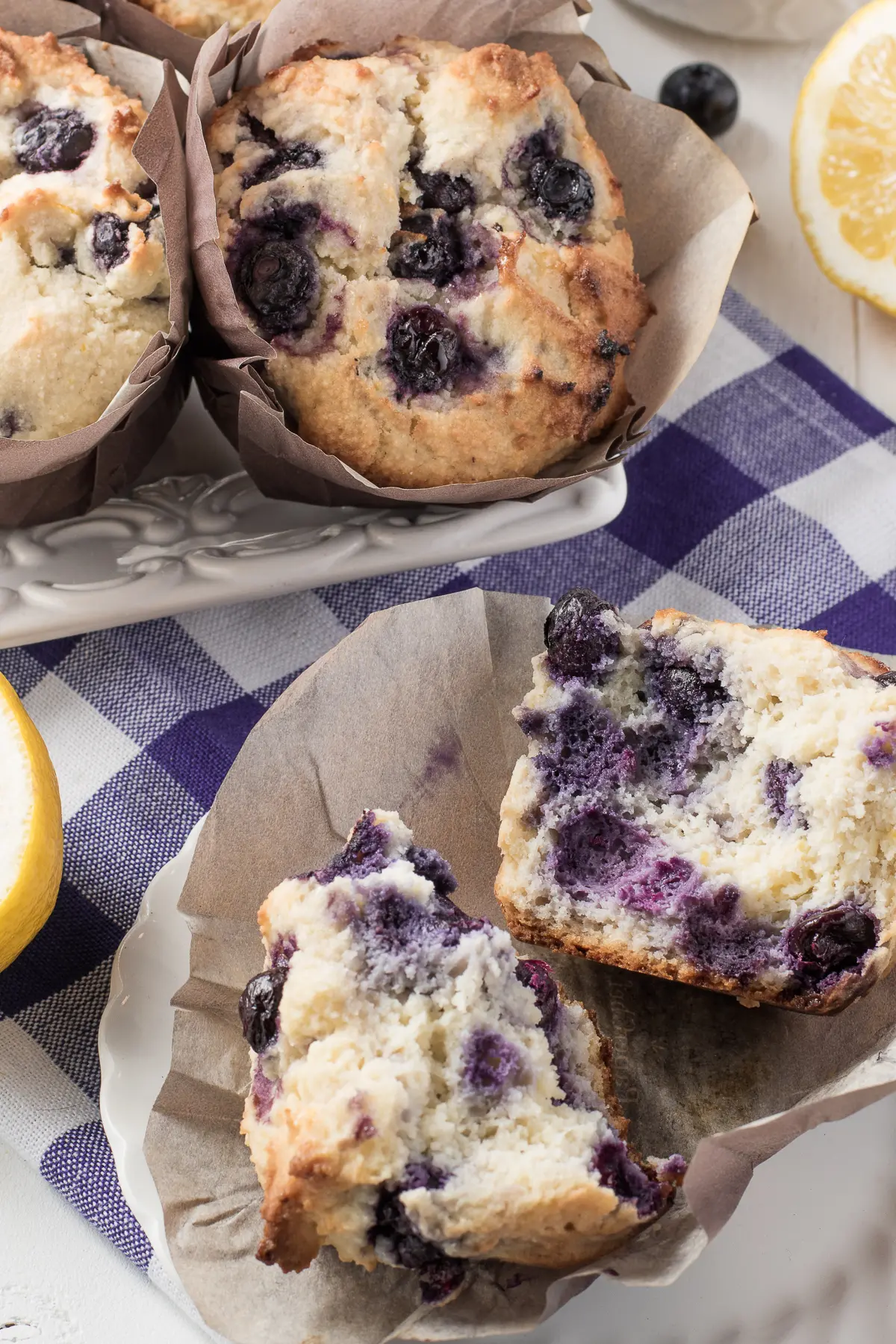 A fresh gluten-free blueberry muffin split down the centre resting in a muffin liner. Surrounded by fresh blueberry muffins and lemons all on a bright blue gingham napkin 