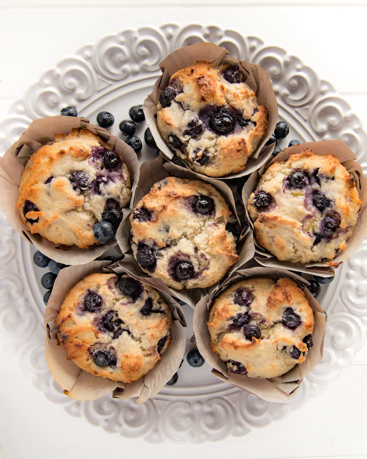A top down shot of low carb blueberry muffins in muffin wrappers laid out on a textured bright white plate. 