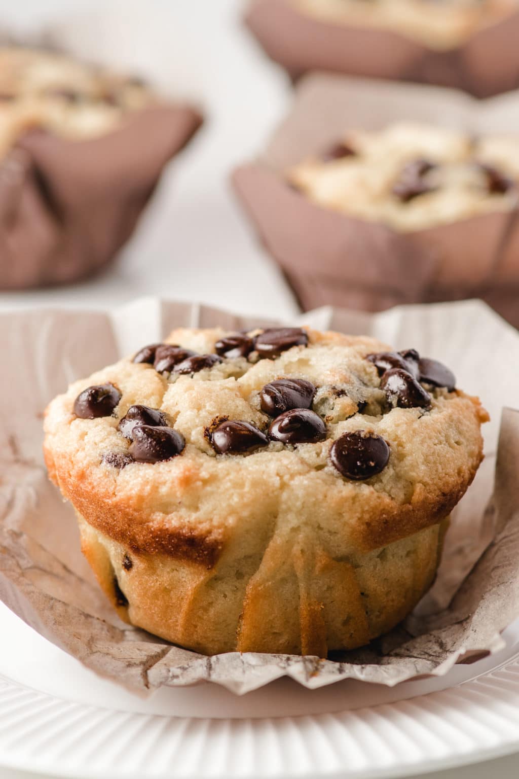 An up close photograph of a bakery style chocolate chip muffin unwrapped on a white plate