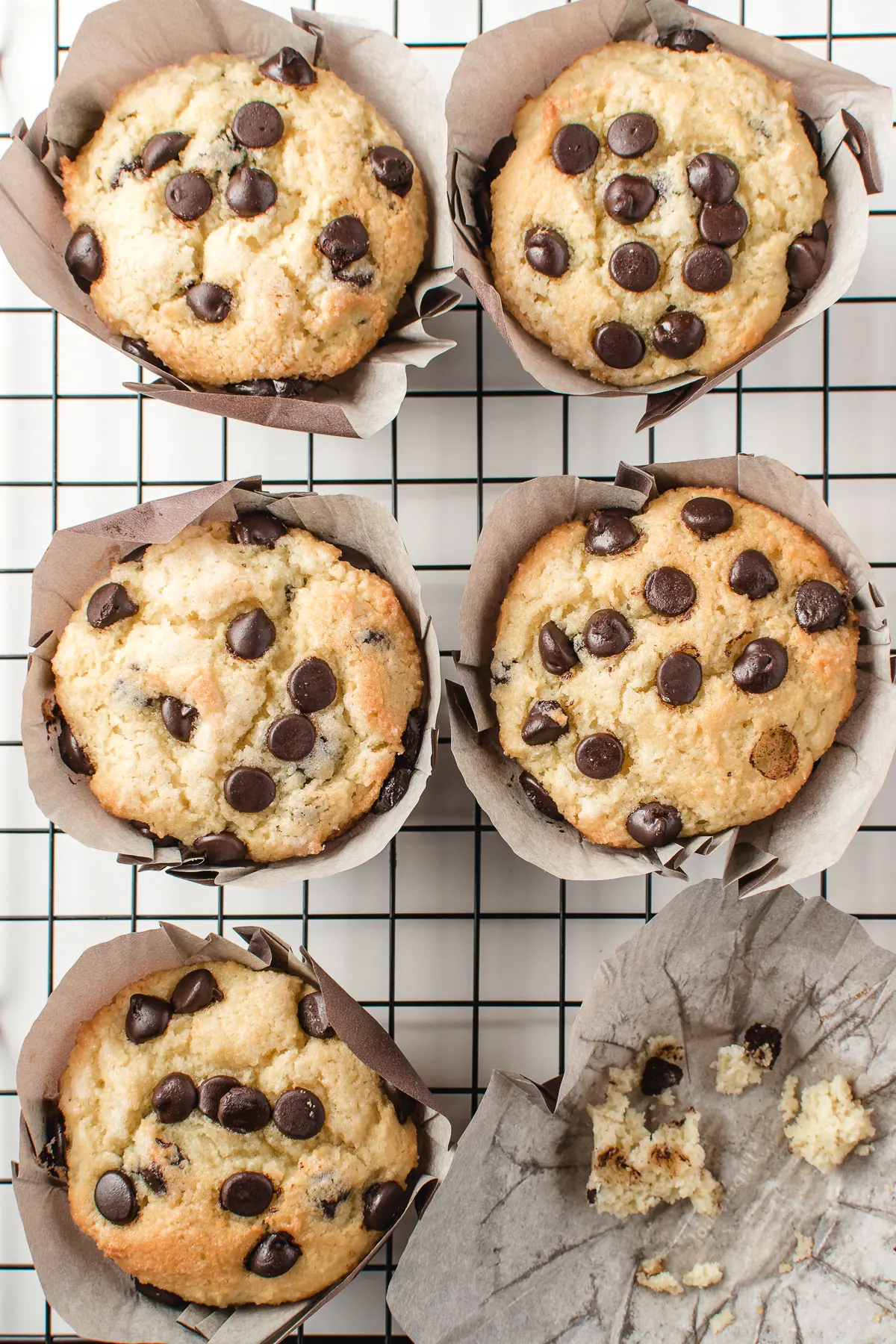 6 bakery style chocolate chip muffins on a black grid cooling rack with one muffin missing leaving only crumbs behind. 