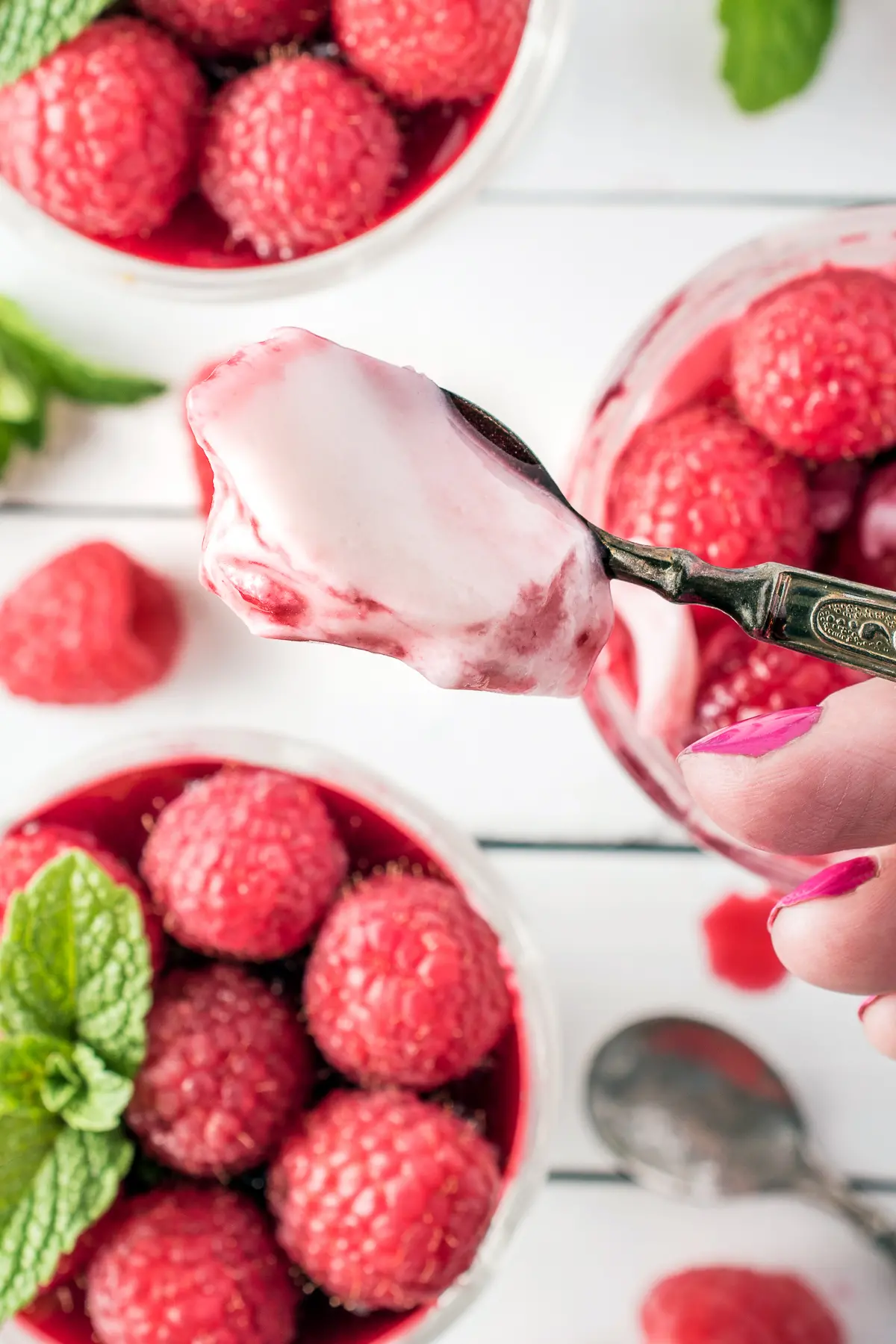 A few fingers are holding a raised teaspoon loaded with raspberry Panna Cotta. The background is white with a few blurred dessert glasses filled with Panna Cotta in the background.