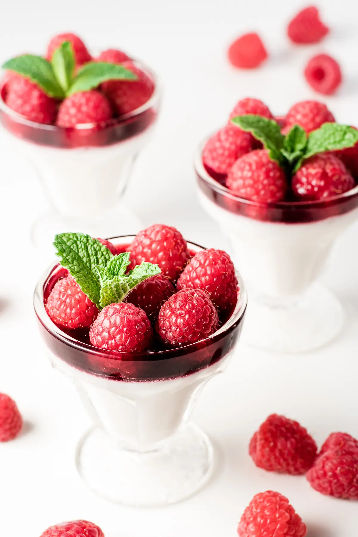 Small sundae dishes with coconut milk raspberry panna cotta topped with red, ripe raspberries and sprigs of mint. There are a few scattered raspberries on the white backdrop.