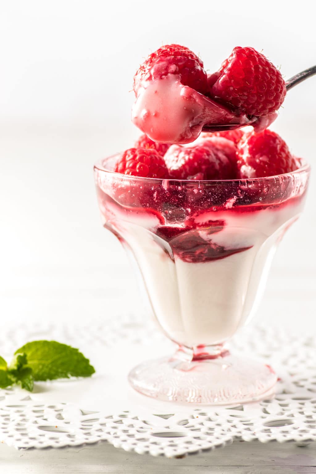 Dessert cup filled with raspberry Panna Cotta. Hovering above the dessert, is a teaspoon loaded with fresh raspberries and Panna Cotta. The dessert cup rests on a decorative white trivet.  A mint leaf  is blurred on the edge of the photo.