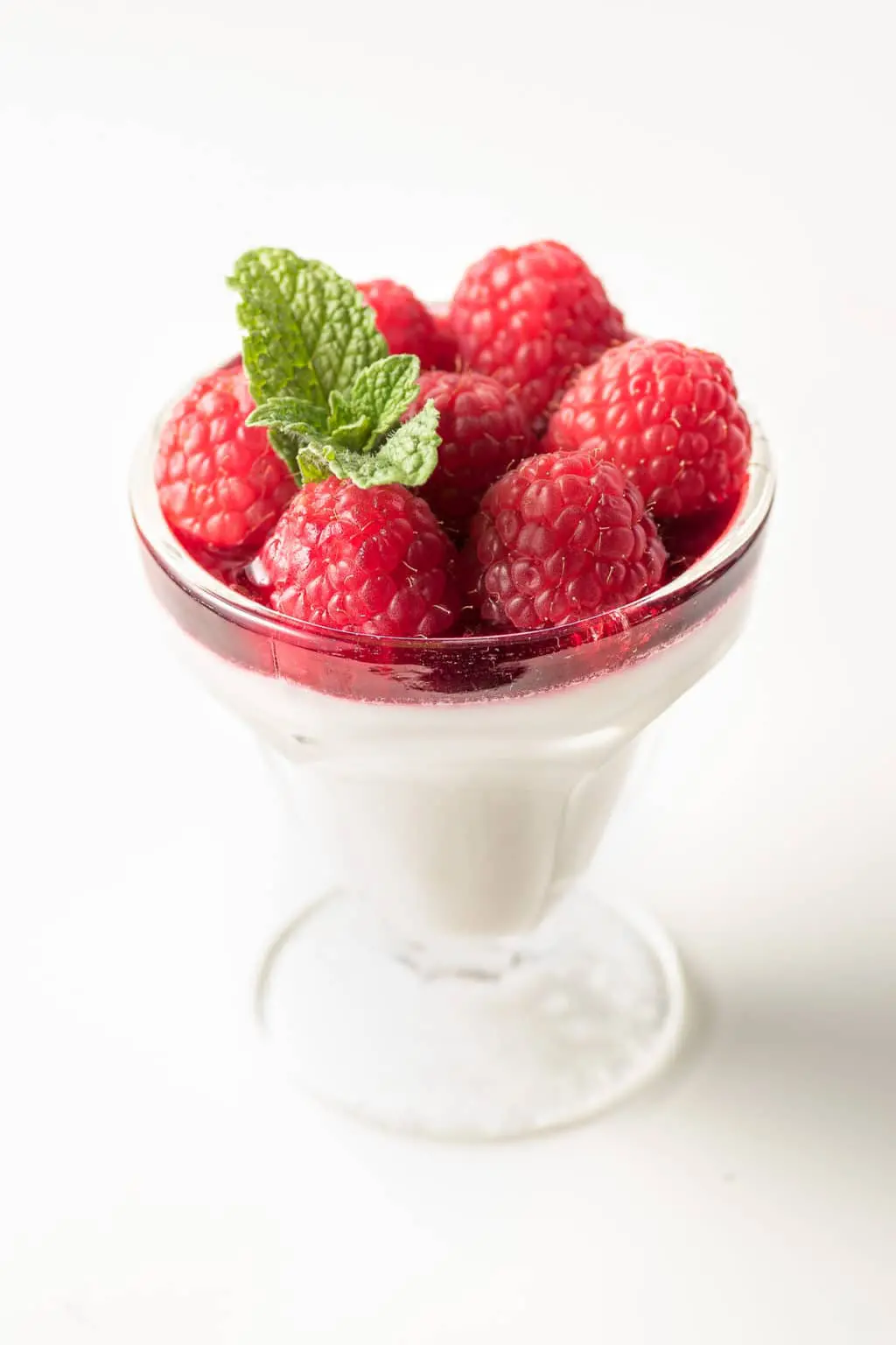 Small sundae dish with coconut milk raspberry panna cotta topped with red ripe raspberries and a sprig of mint.