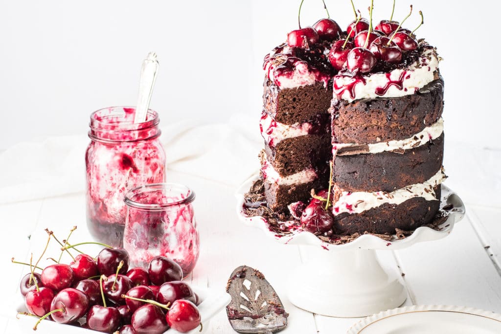 A three layer dark chocolate low carb black forrest cake, filled and topped with vanilla whipped cream frosting and drizzled with cherry syrup on a bright white table top. 