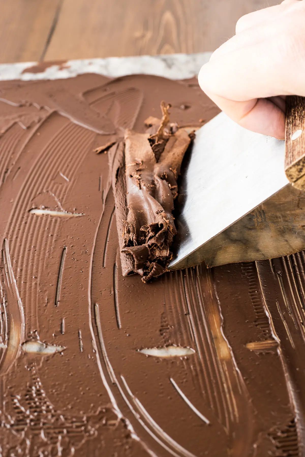 Melted chocolate on a marble board being pushed with a pastry scraper to create large flakes and ribbons of dark chocolate