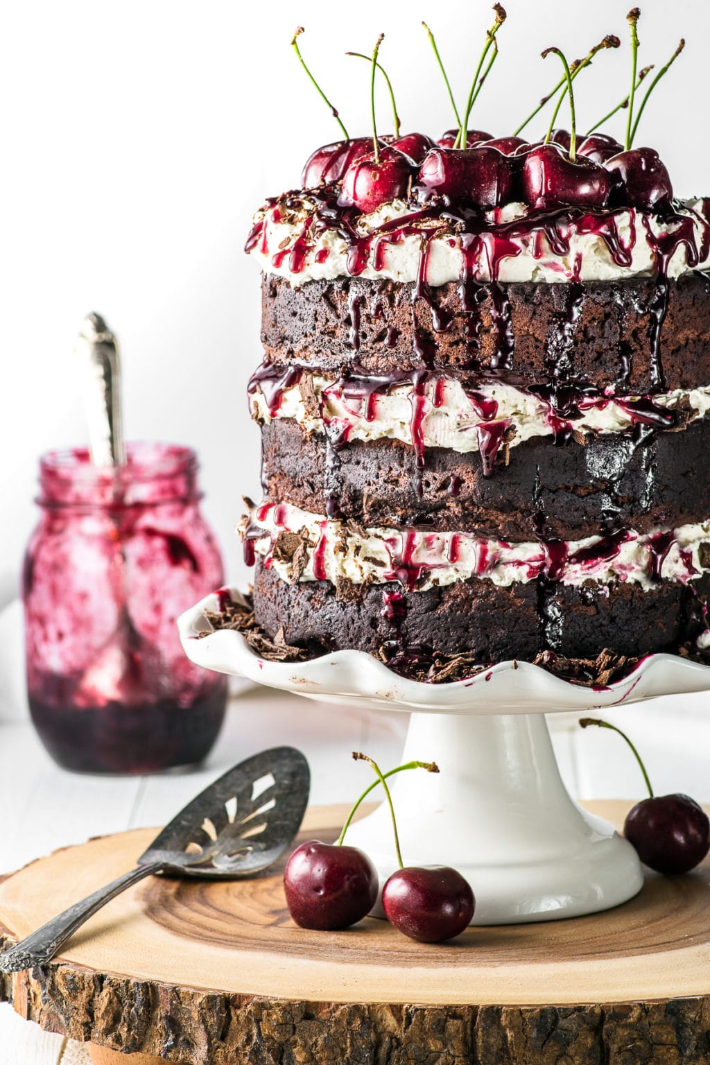 A tall triple layer gluten-free black forest cake topped with whipped cream frosting and fresh cherries dripping cherry filling against a white background