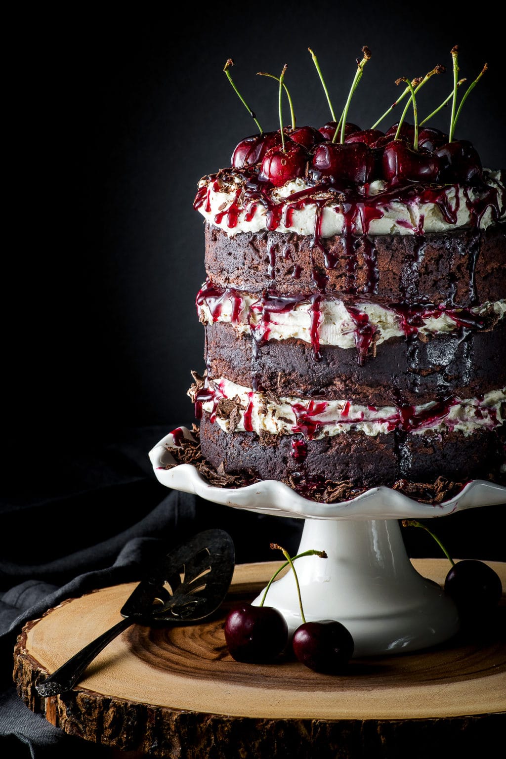 Black forest cake on a white cake stand, covered in fresh cherries against a dark black background.