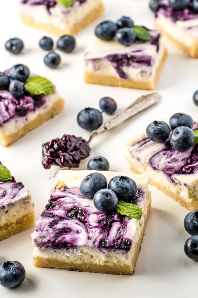 Blueberry cheesecake bars topped with swirls of pureed blueberries, fresh blueberries, and sprigs of mint. A teaspoon filled with sugar-free blueberry jam sits in the centre.