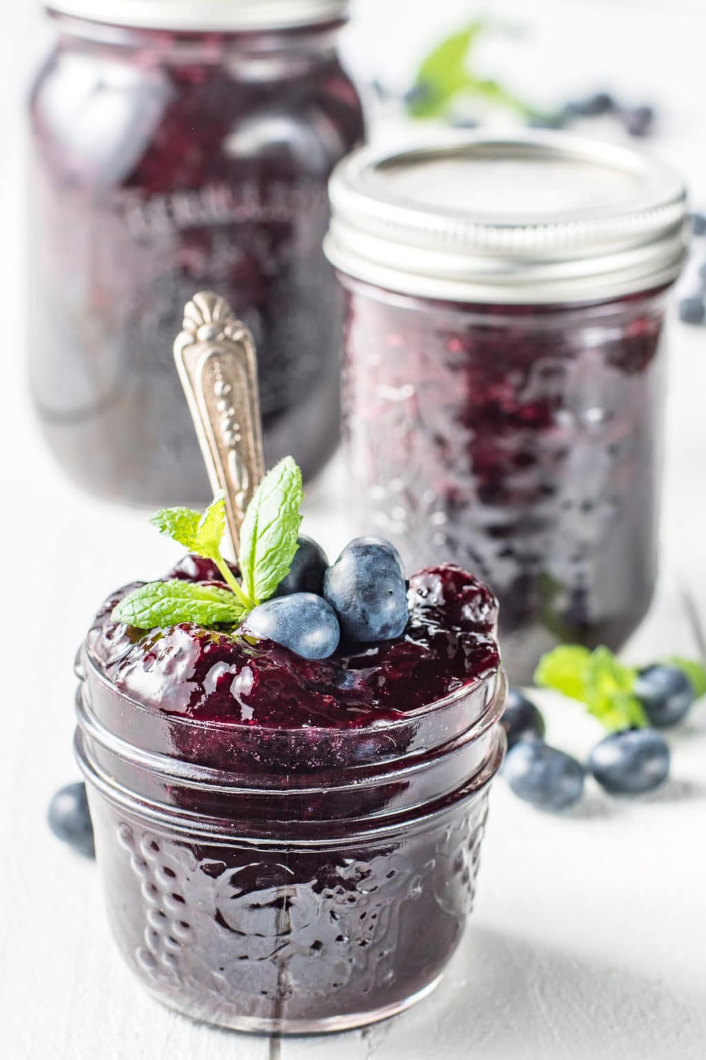 Jars of sugar-free blueberry jam on a white wooden board with sprigs of mint and scattered fresh blueberries