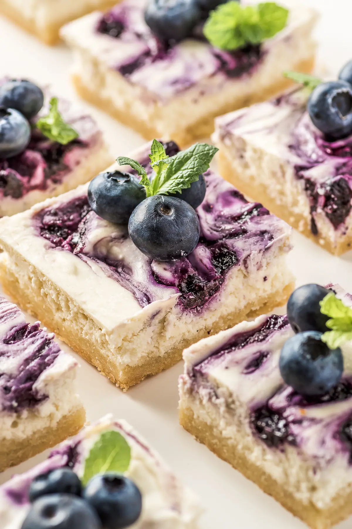 Close-up photo of keto blueberry cheesecake bars set on a white plate. They are topped with fresh blueberries and mint sprigs.