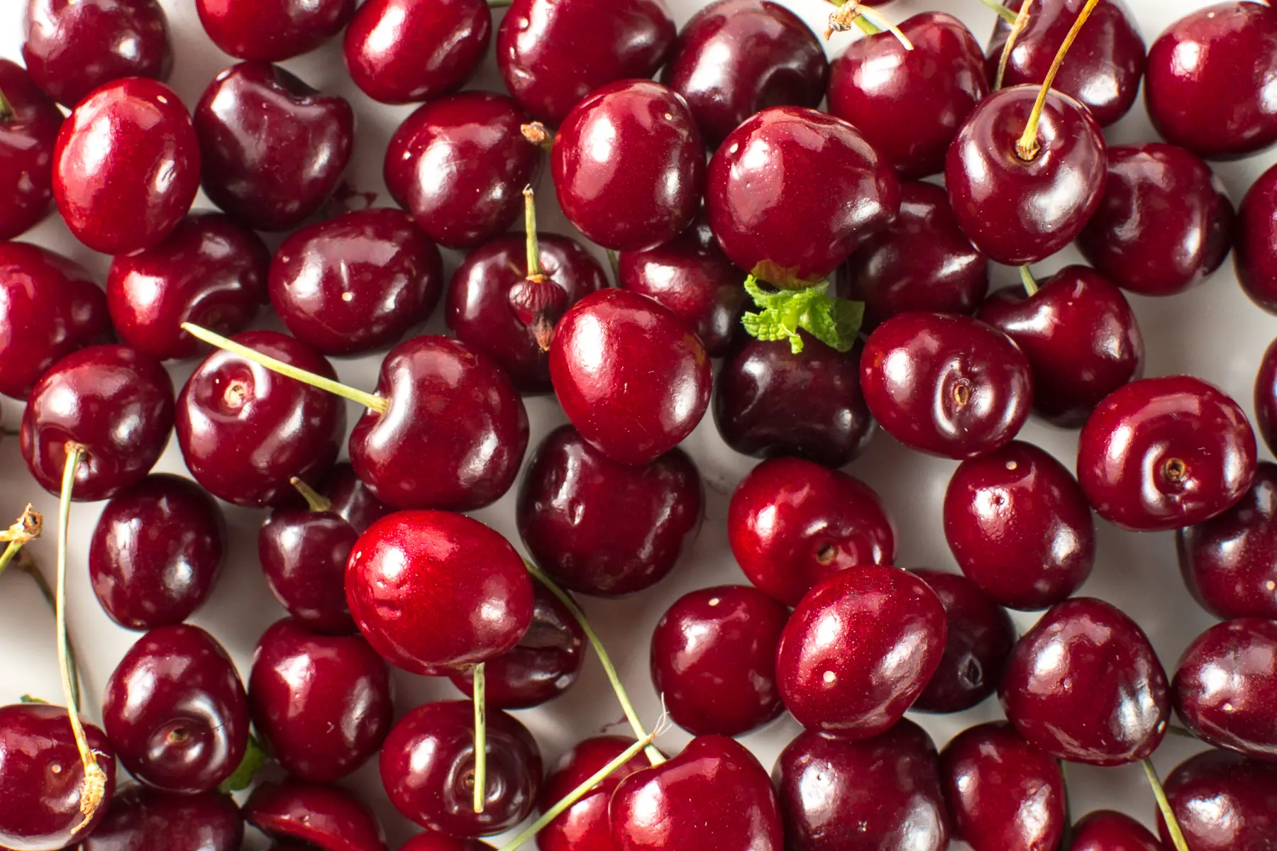 A close up photograph of a bright red fresh stemmed cherries on a bright white table top. 