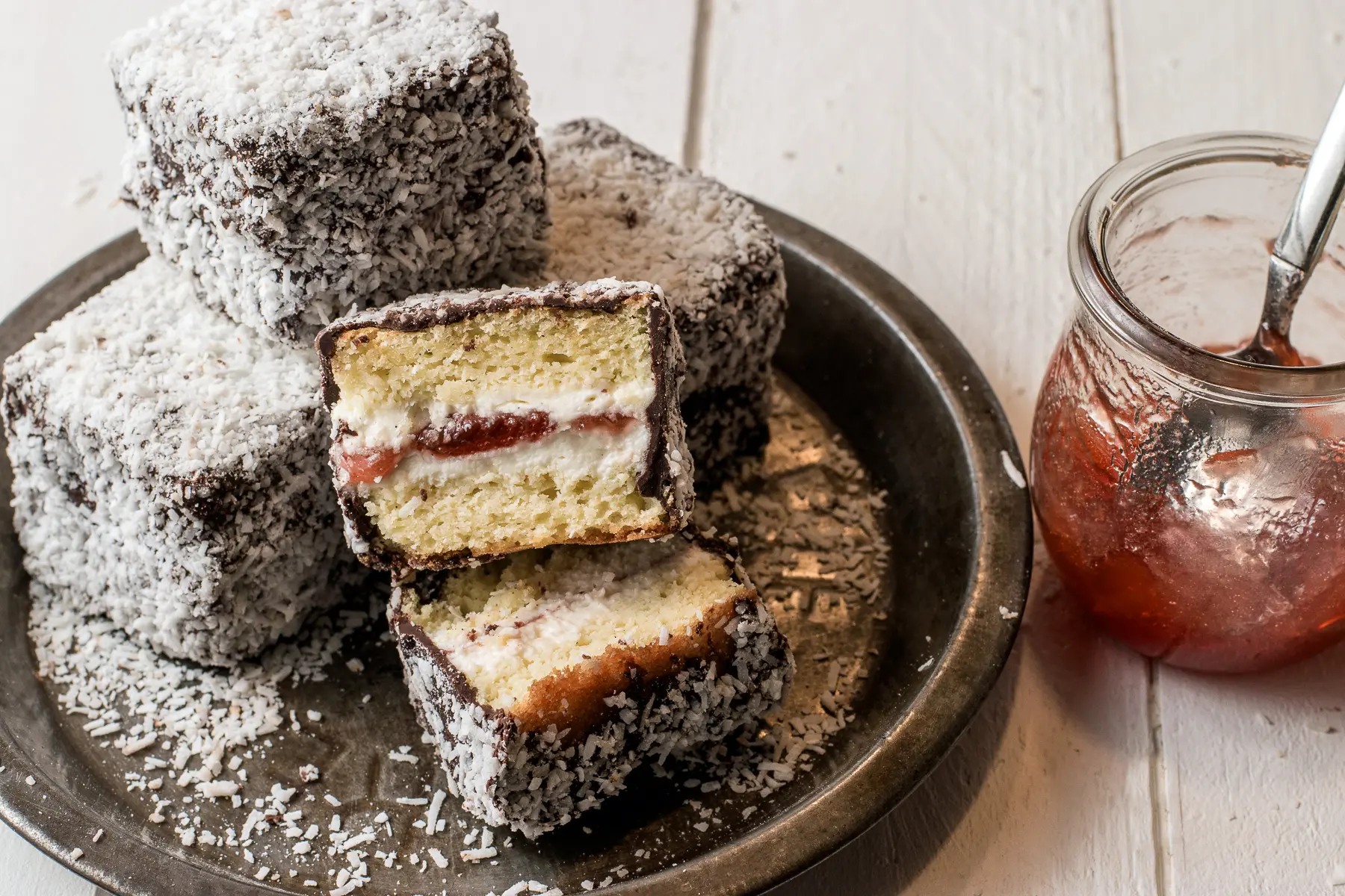 Gluten-free jam filled lamingtons on a rustic pie tin with a jar of strawberry jam.