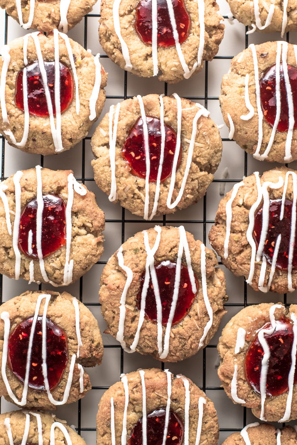 Resting on a white backdrop, is a metal cookie rack covered with raspberry jam-filled, thumbprint cookies drizzled with sugar-free, white glaze.