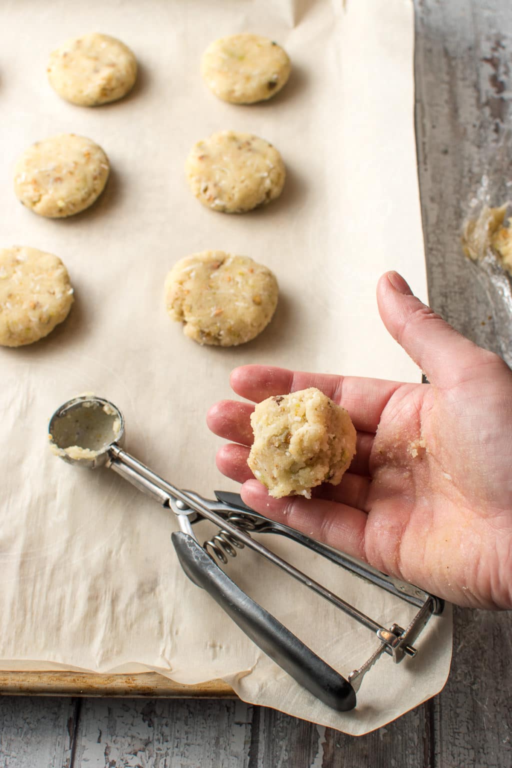 There are six, flattened balls of cookie dough spaced evenly on a piece of parchment paper. On the right, is a person's open hand, holding a ball of cookie dough. Resting on the parchment in the lower left, is an empty, stainless cookie scoop with a bit of cookie dough stuck to the inside of it.