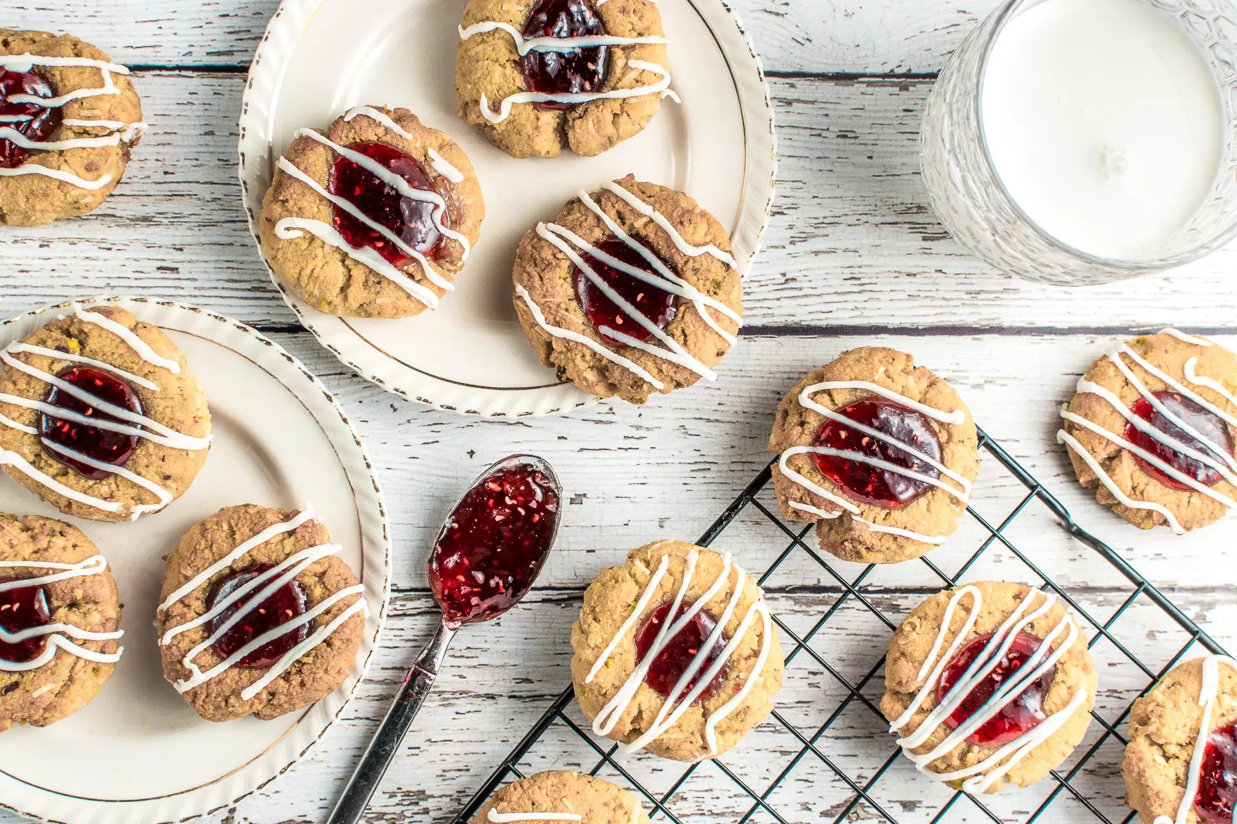 Keto raspberry jam thumbprint cookies on two, ivory, vintage dessert plates set on a rustic white barnyard backdrop. A glass of milk and a cooling rack with more cookies, along with a teaspoon filled with gooey, raspberry jam, complete the picture.