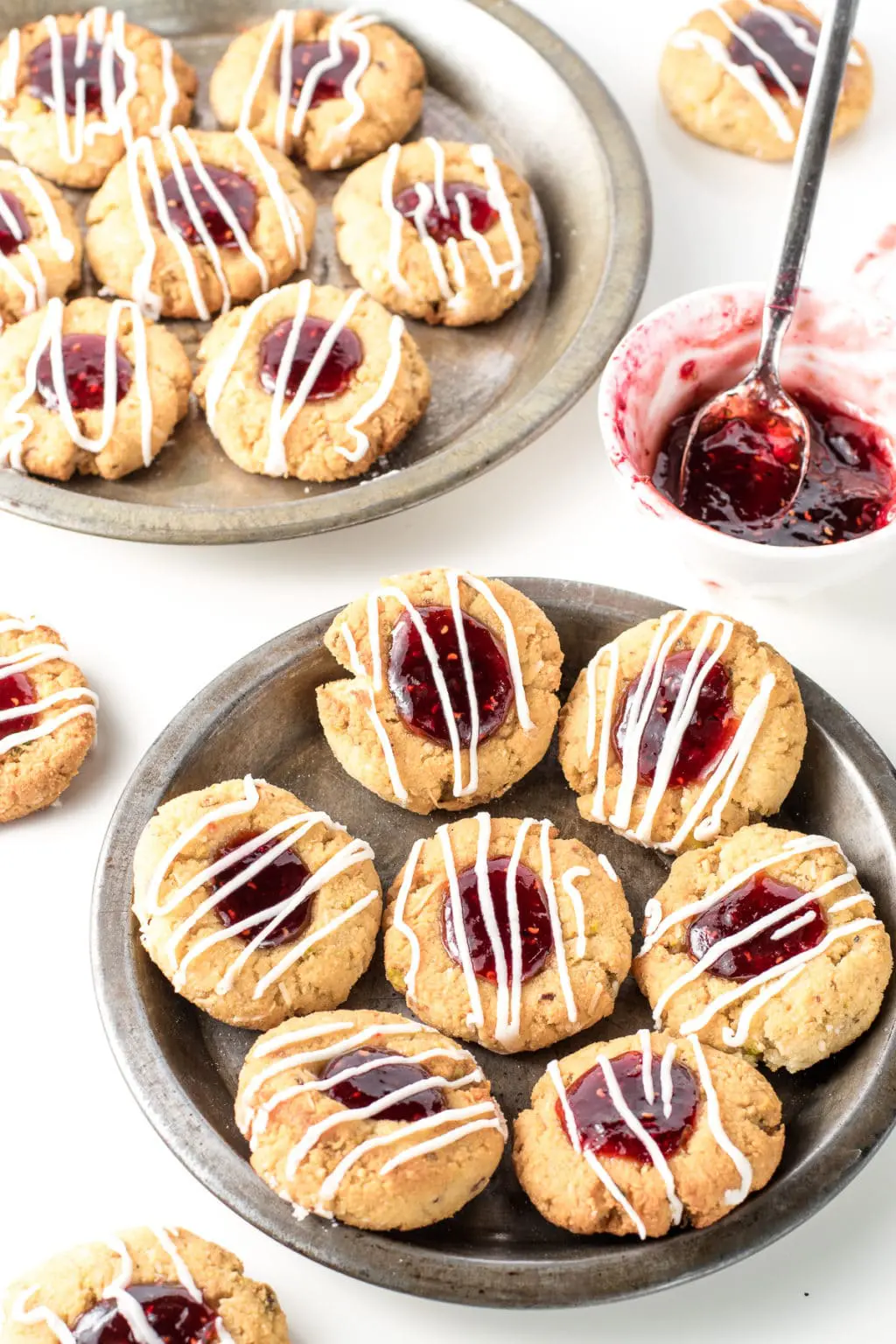 Two metal pie plates filled with thumbprint cookies. There are two cookies resting directly on the white background. To the right of centre is a small, white bowl with a teaspoon in it.  They are filled with gooey, raspberry jam.