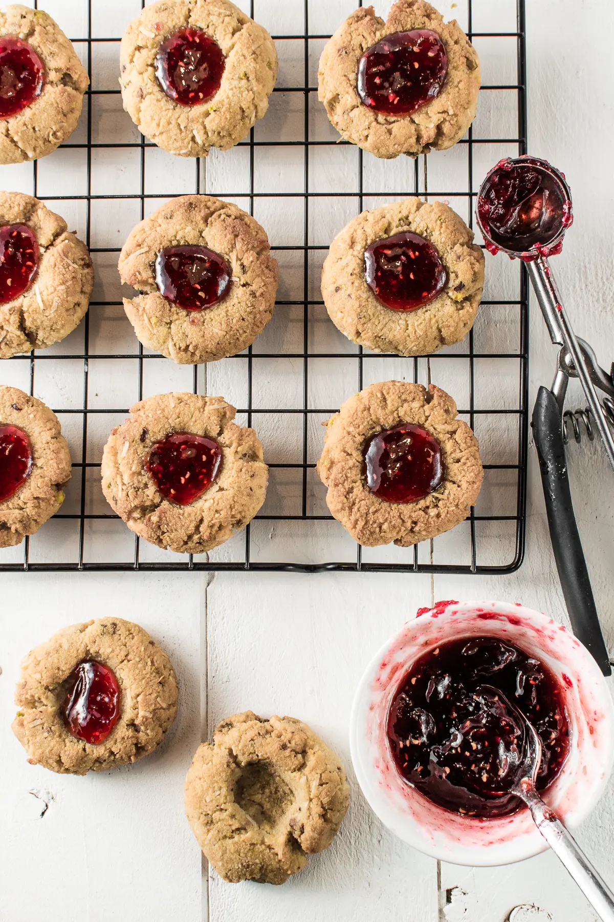 Set on a white backdrop, are nine thumbprint cookies filled with gooey, raspberry jam spaced evenly on a cooling rack. A stainless cookie scoop, half full of sticky jam, rests on the right side of the metal rack.  Below the rack, are two cookies--one filled with jam, and one with an empty divot. Beside the empty cookie is a jam-filled teaspoon inside a small, white bowl filled with sticky, raspberry jam.