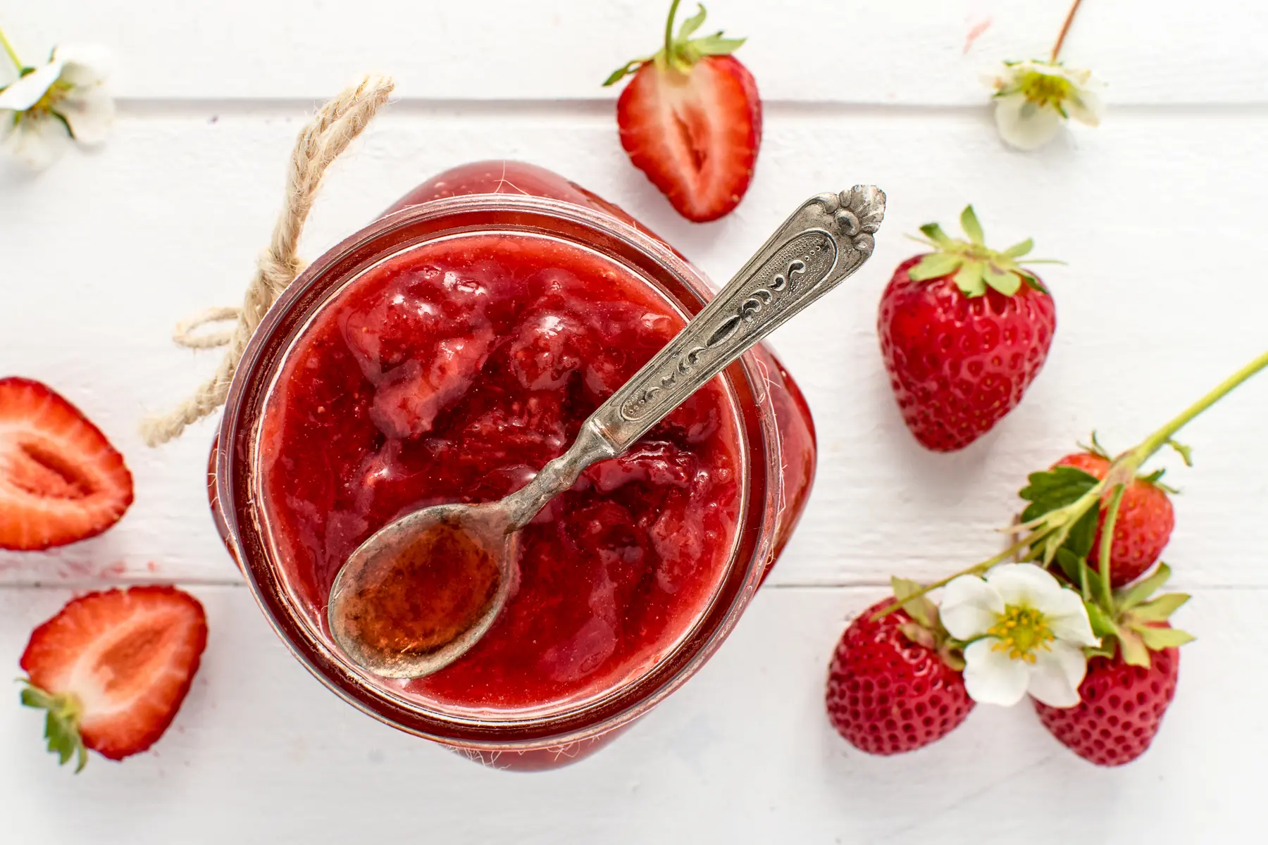 Top down photo of an open jar of sugar-free strawberry jam with fresh strawberries and flowers scattered . 