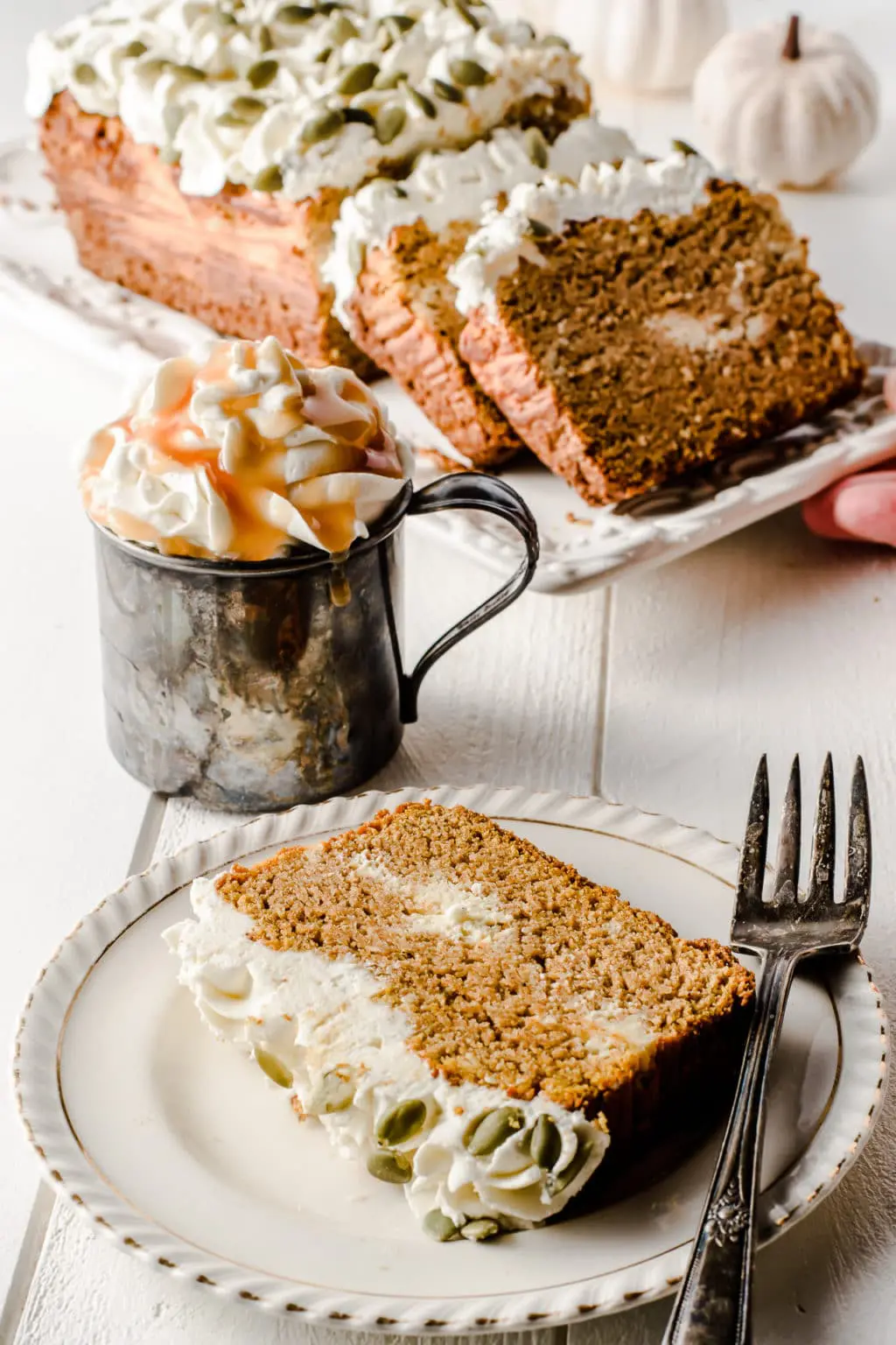 A slice of low carb pumpkin bread on a plate, with a hot beverage loaded with whipped cream and caramel sauce.