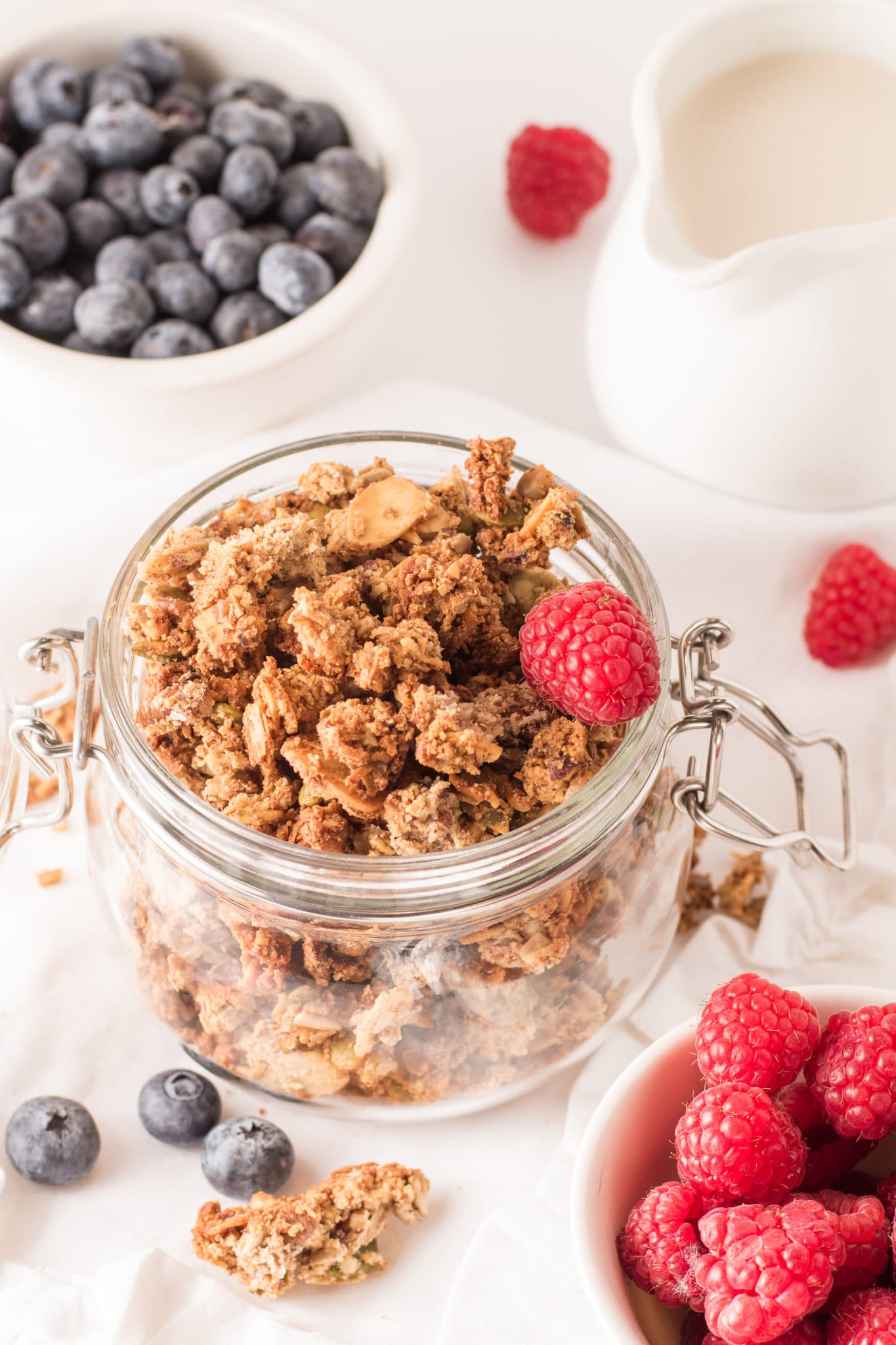 A glass mason jar filled with low carb granola cluster surrounded by bowls of fresh blueberries and raspberries. 