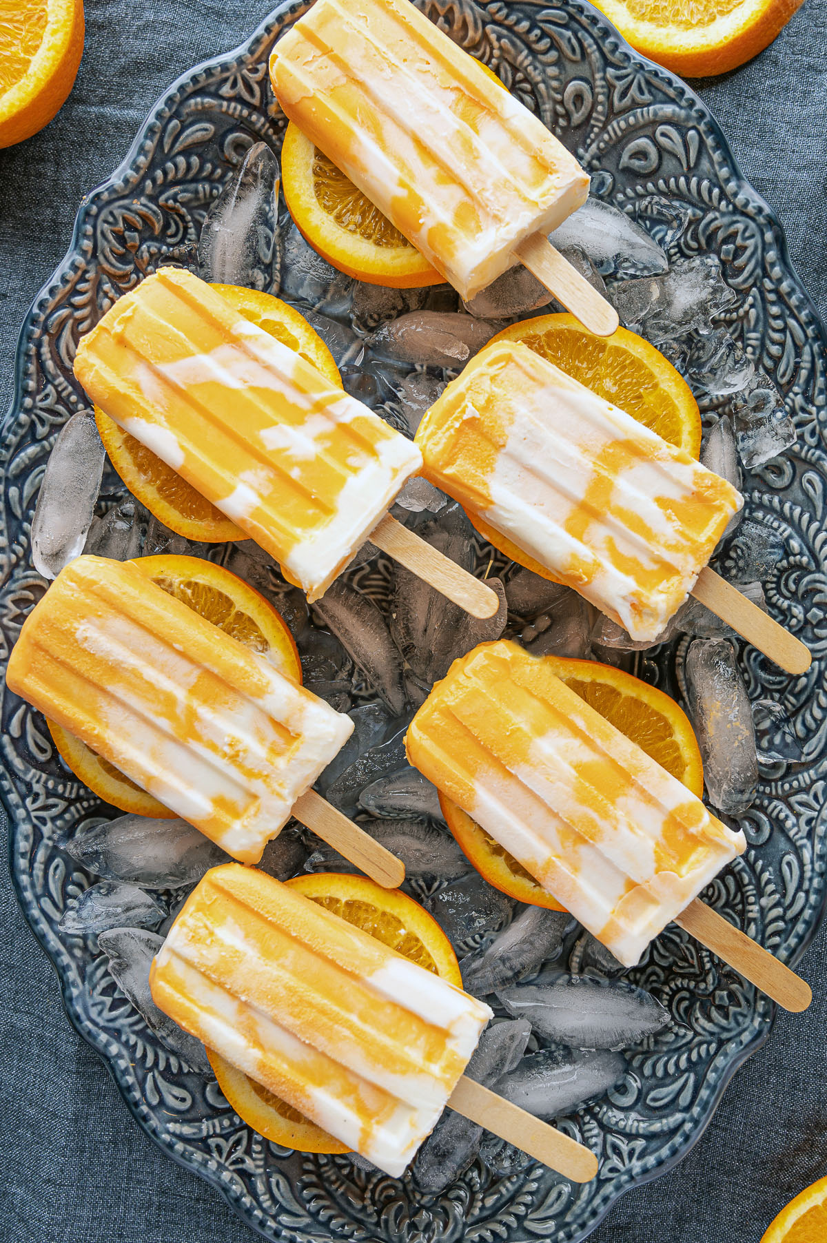 Orange and white marbled creamsicle popsicles resting on a blue platter of ice and orange slices