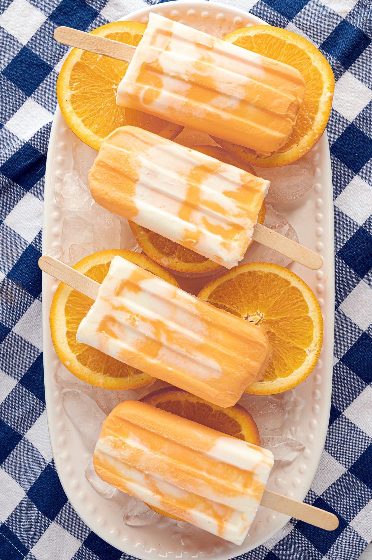 Orange and white marbled creamsicle popsicles resting on a white platter on top of a blue gingham table cloth