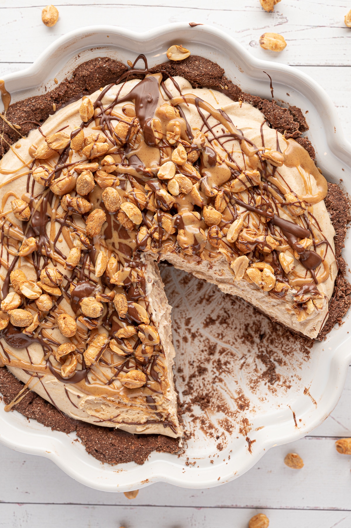 A chocolate crusted peanut butter pie in a white pie pan, peanuts, chocolate and peanut butter drizzled over the top, with slice removed. 