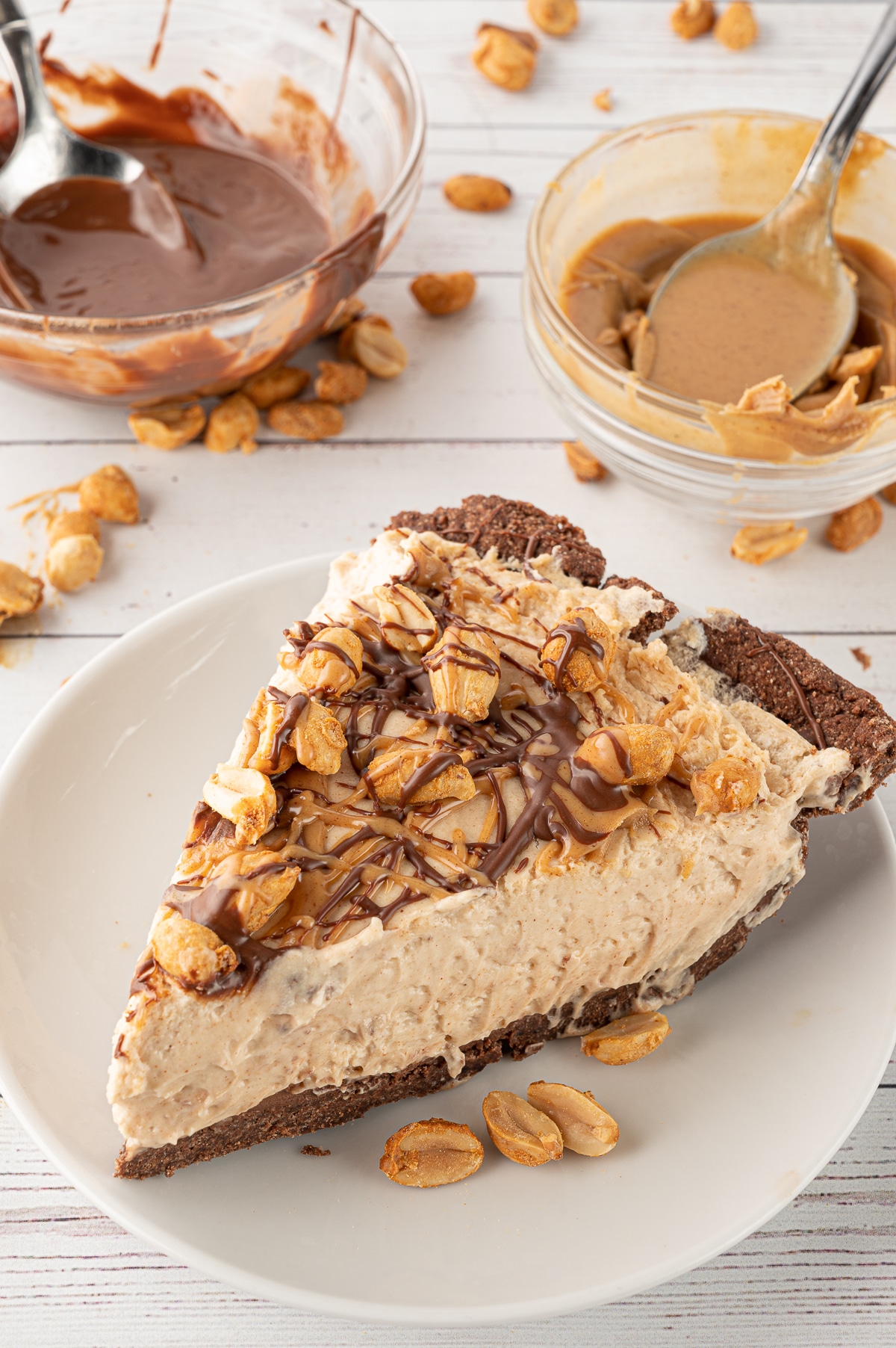 A slice of peanut butter pie with scattered peanuts, and bowls of melted chocolate ganache and peanut butter in the background