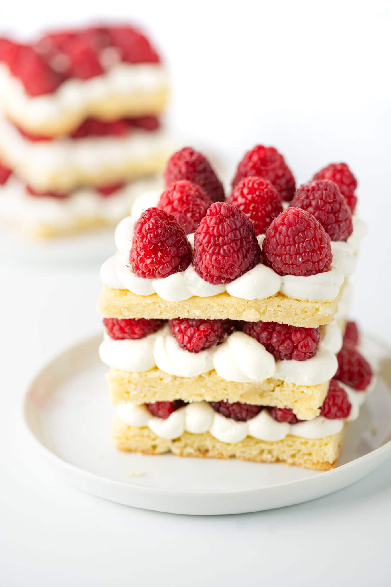 A three layer square short cake with bright fresh red raspberries,and whipped cream filling on a small white plate. 