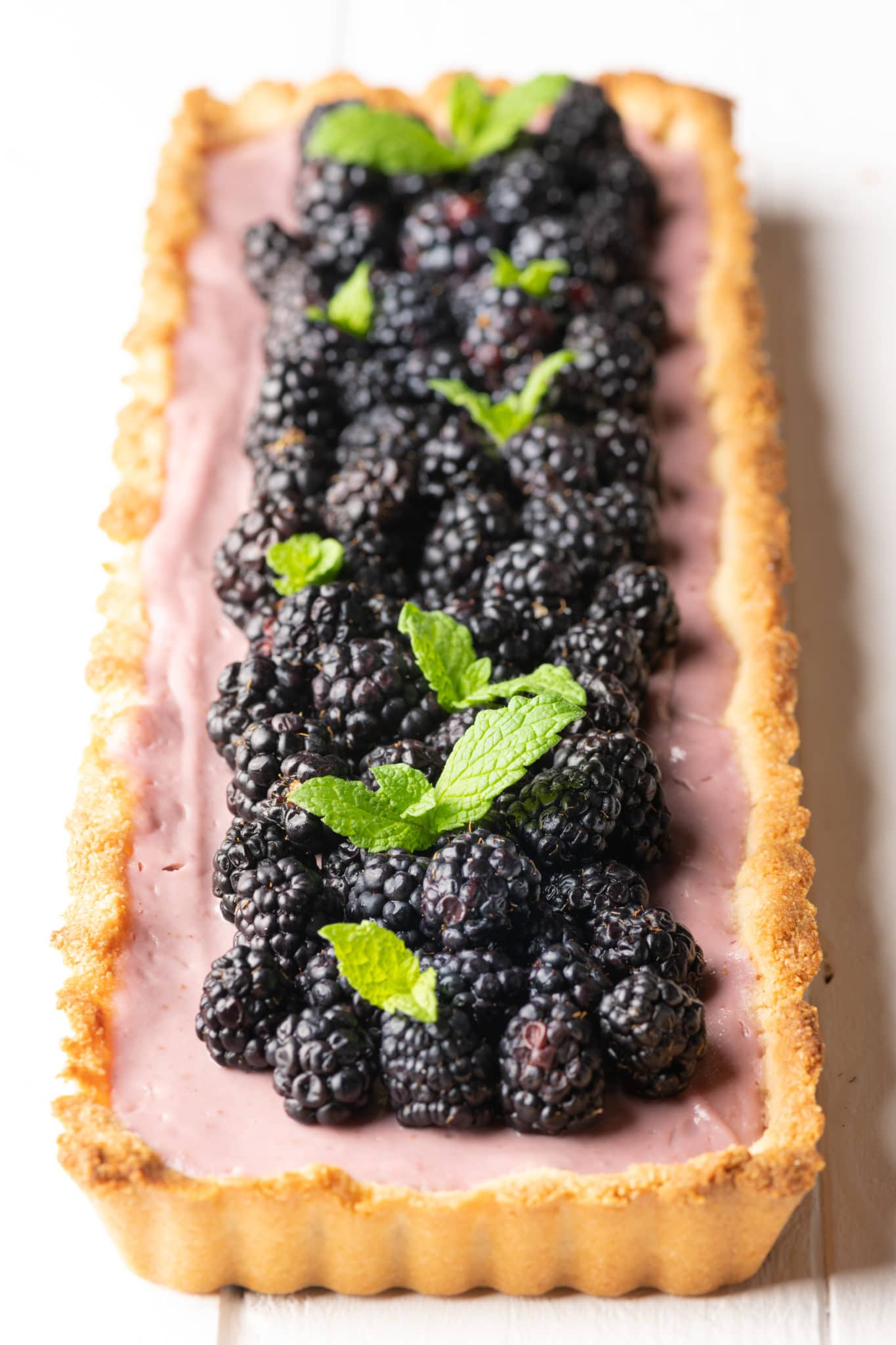 Rectangular shaped tart with golden crust and light purple filling topped with fresh blackberries and sprigs of mint. 