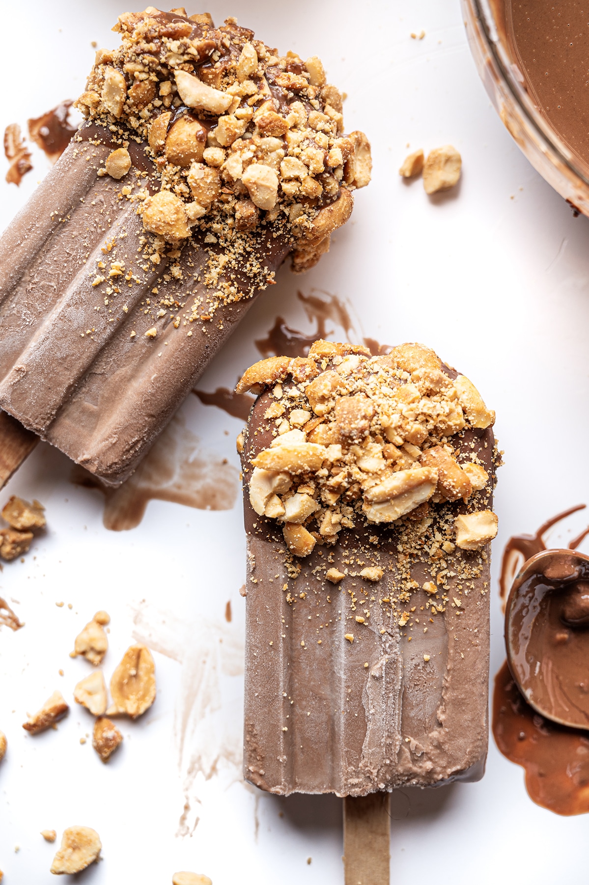 Frozen sugar free fudgesicles melting on a bright white background with smeared chocolate spoon and scattered crushed peanuts