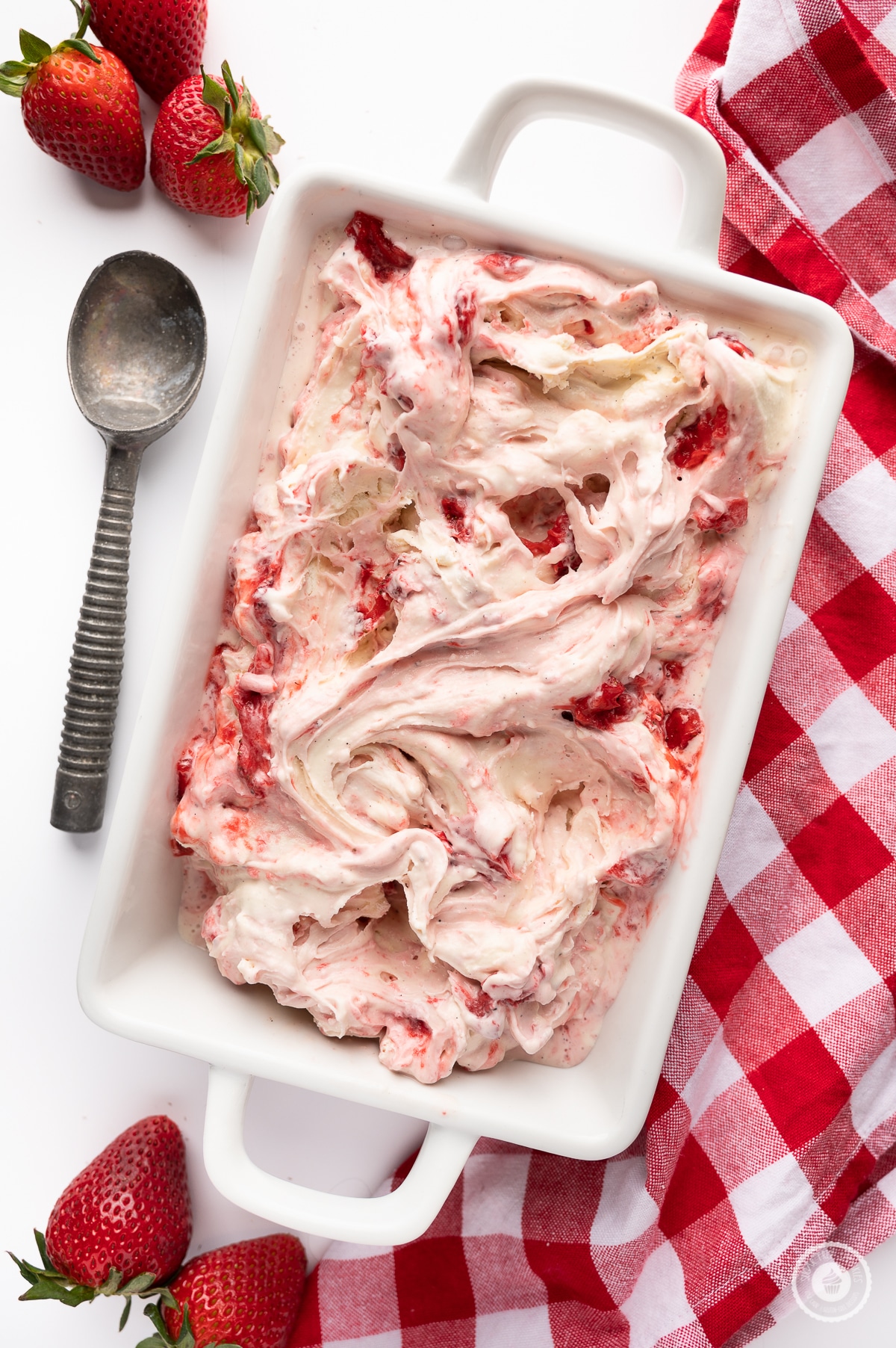 Low carb strawberry cheesecake ice cream in a ceramic dish with a vintage ice cream scoop, fresh berries and red and white check napkin all on a bright white background