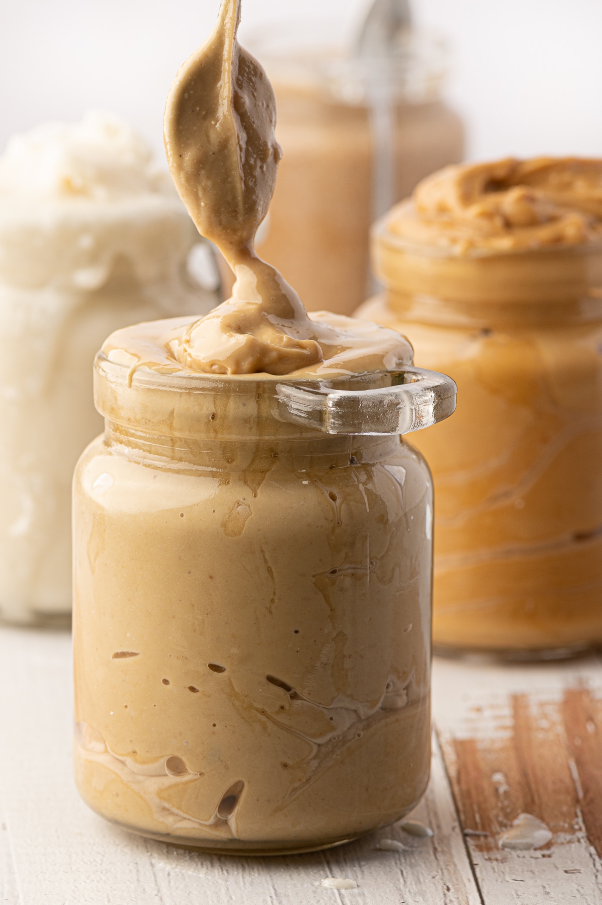 A jar of cashew butter being spooned into a glass jar with jars of overfull nut butters in the background 