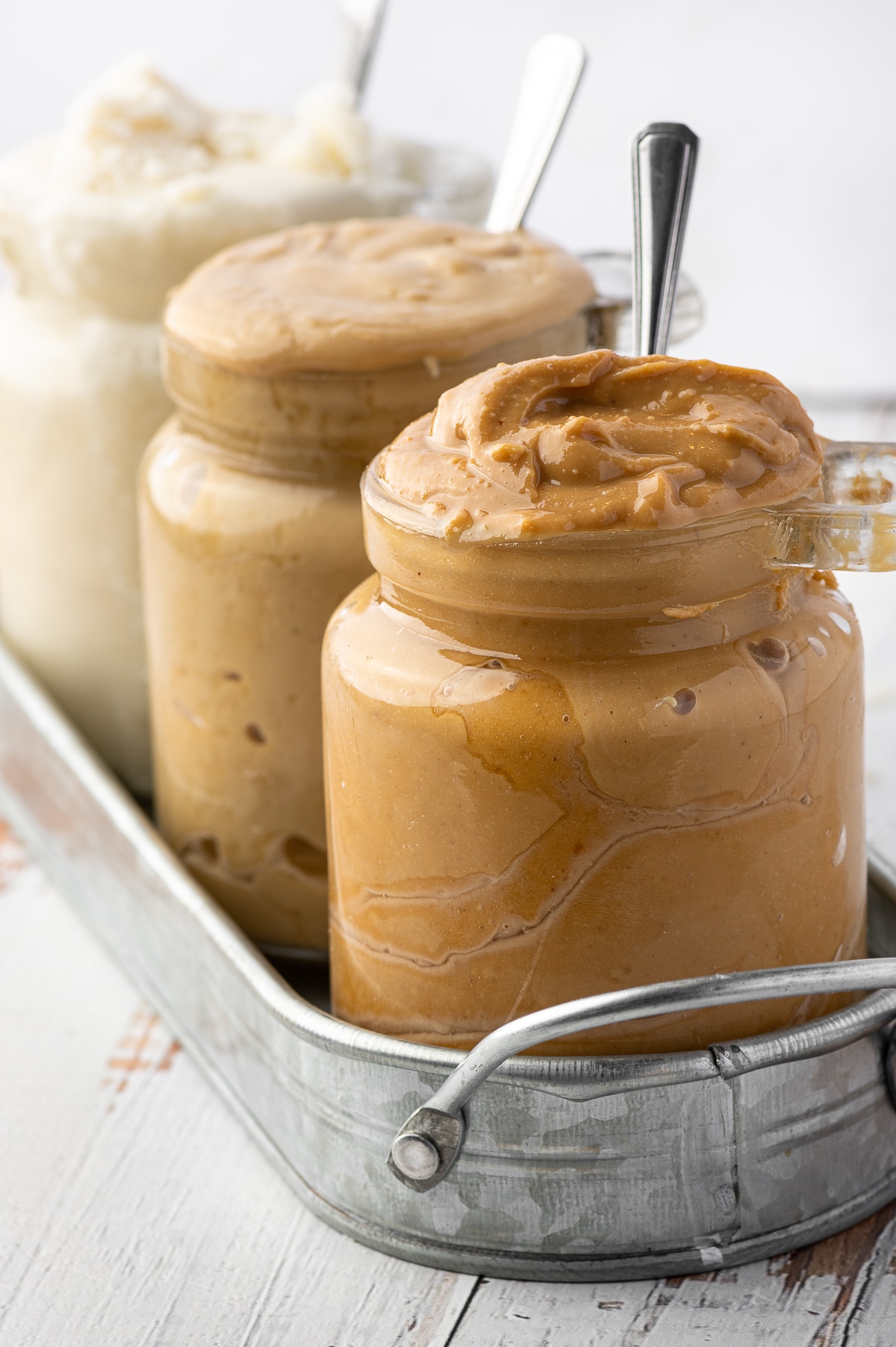 Three overflowing jars of nut butters in a metal jar against a bright white background 